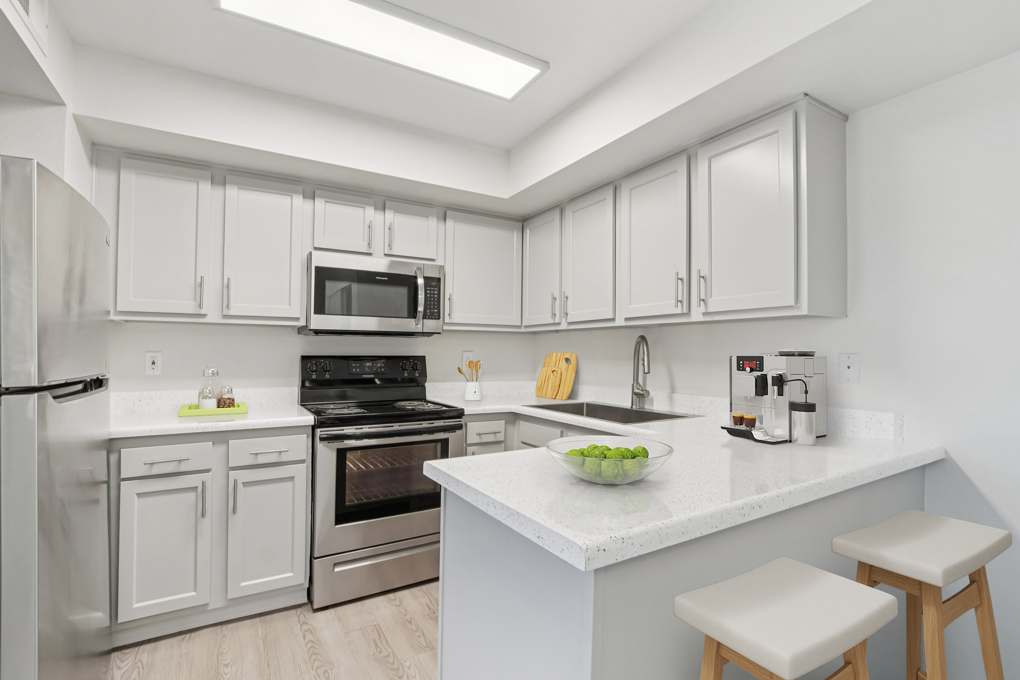 Modern kitchen featuring gray cabinetry, stainless steel appliances including a microwave and oven, a white countertop with a bowl of limes, and two wooden stools. Brightly lit with natural light, the space is organized and inviting, showcasing a contemporary design with sleek finishes.