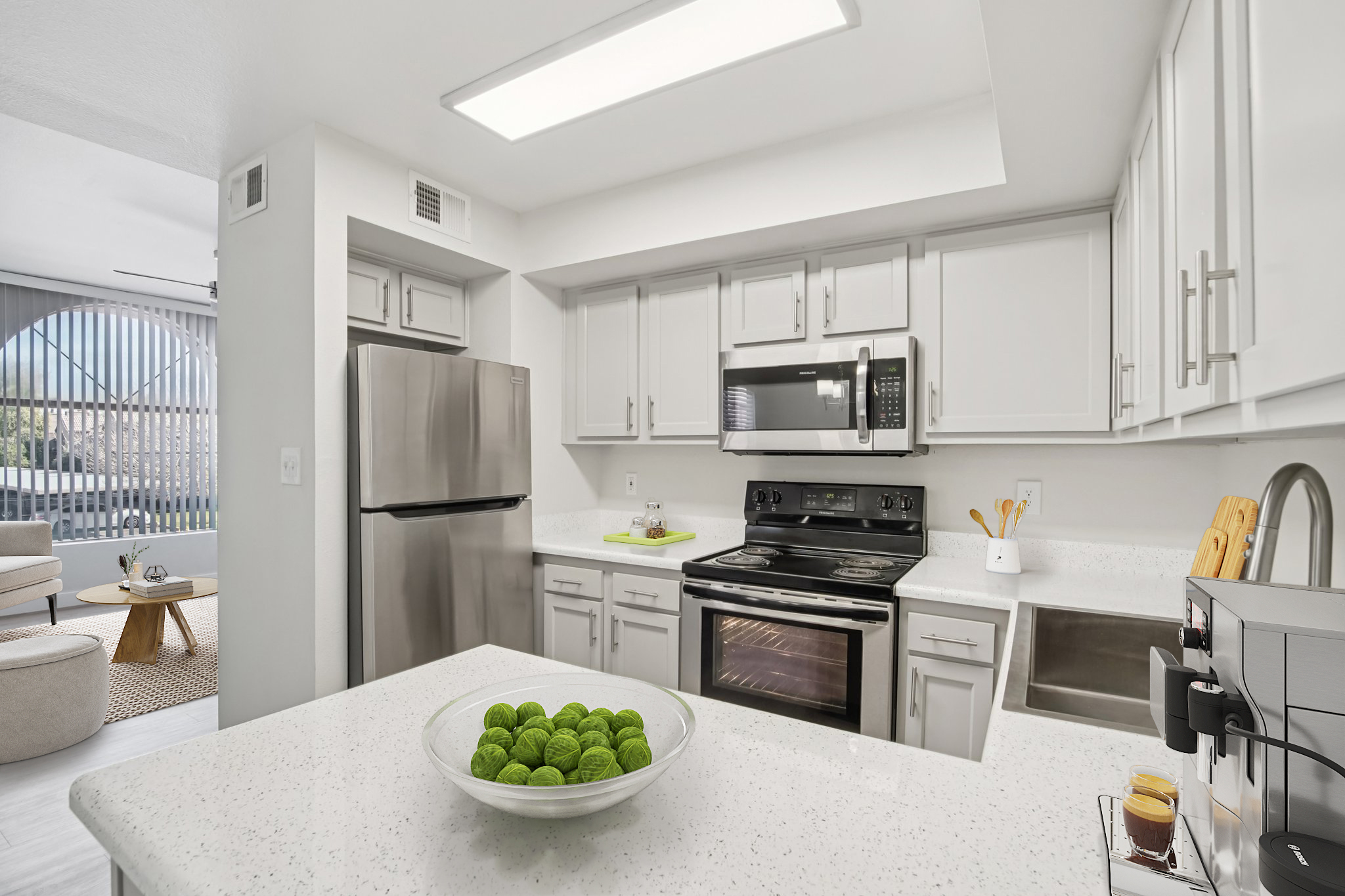 Modern kitchen with stainless steel appliances, including a refrigerator and stove. The countertops are white with a bowl of green decorative spheres. The cabinetry is light-colored, and a window can be seen in the background, suggesting a bright, open space.