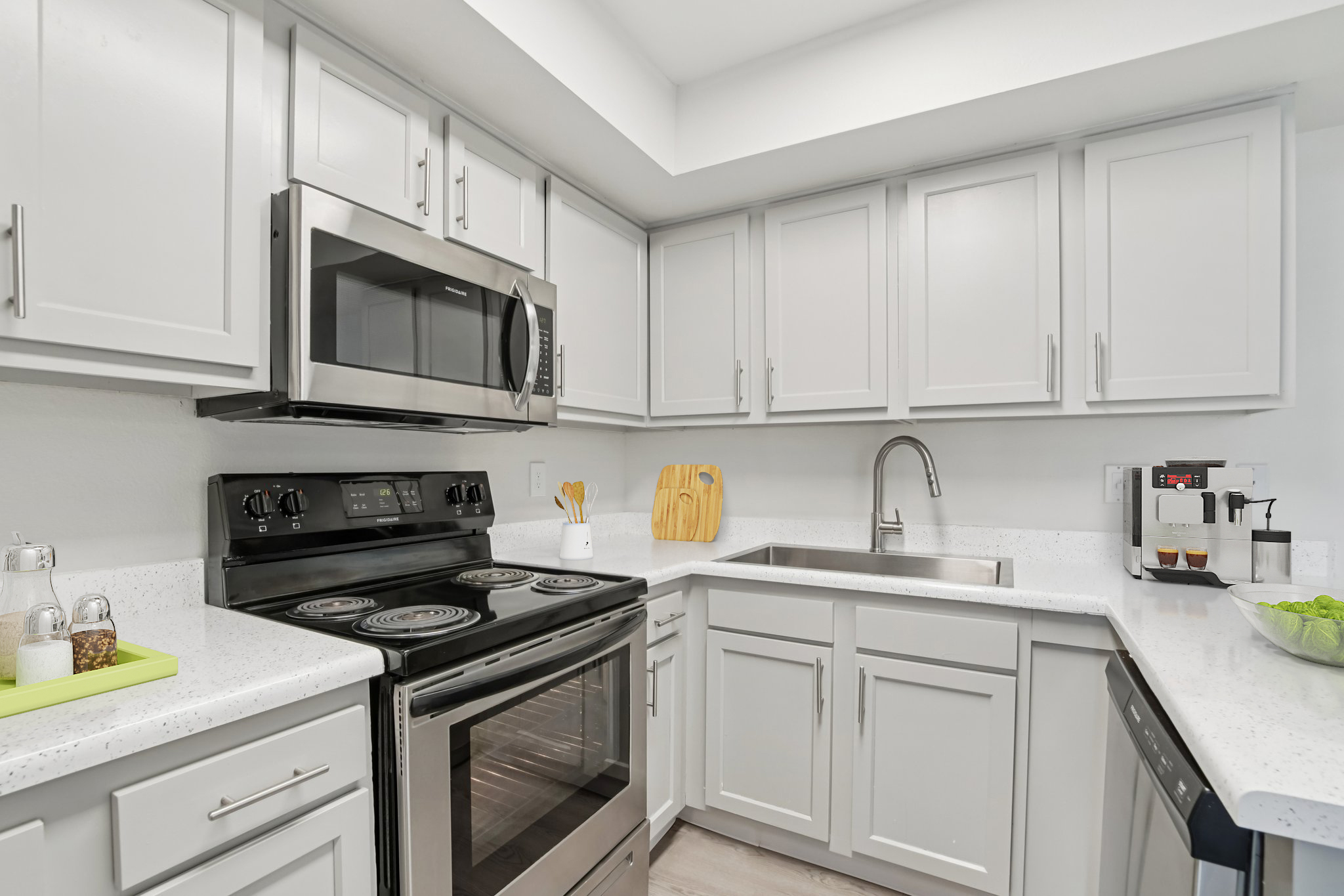 Modern kitchen featuring stainless steel appliances, including a microwave and oven, set against light grey cabinetry. A spacious sink is positioned next to a coffee maker and cutting board on the counter. The design emphasizes a clean, bright aesthetic with a neutral color palette and no visible clutter.
