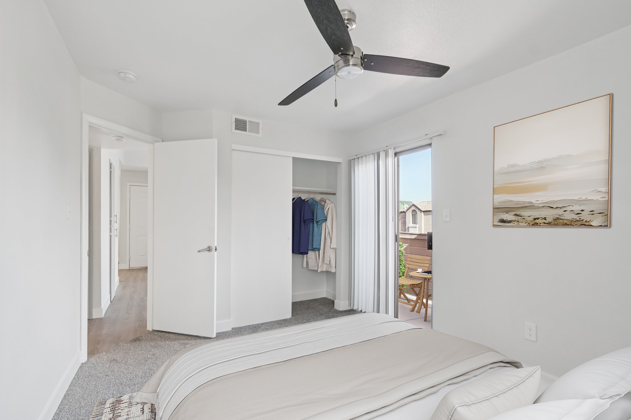 A bright, modern bedroom featuring a neatly made bed with a beige throw blanket, a ceiling fan, and light gray walls. A closet is visible with clothes hanging. There is a sliding glass door leading to a small balcony with outdoor furniture, and a framed artwork of a landscape is on the wall.