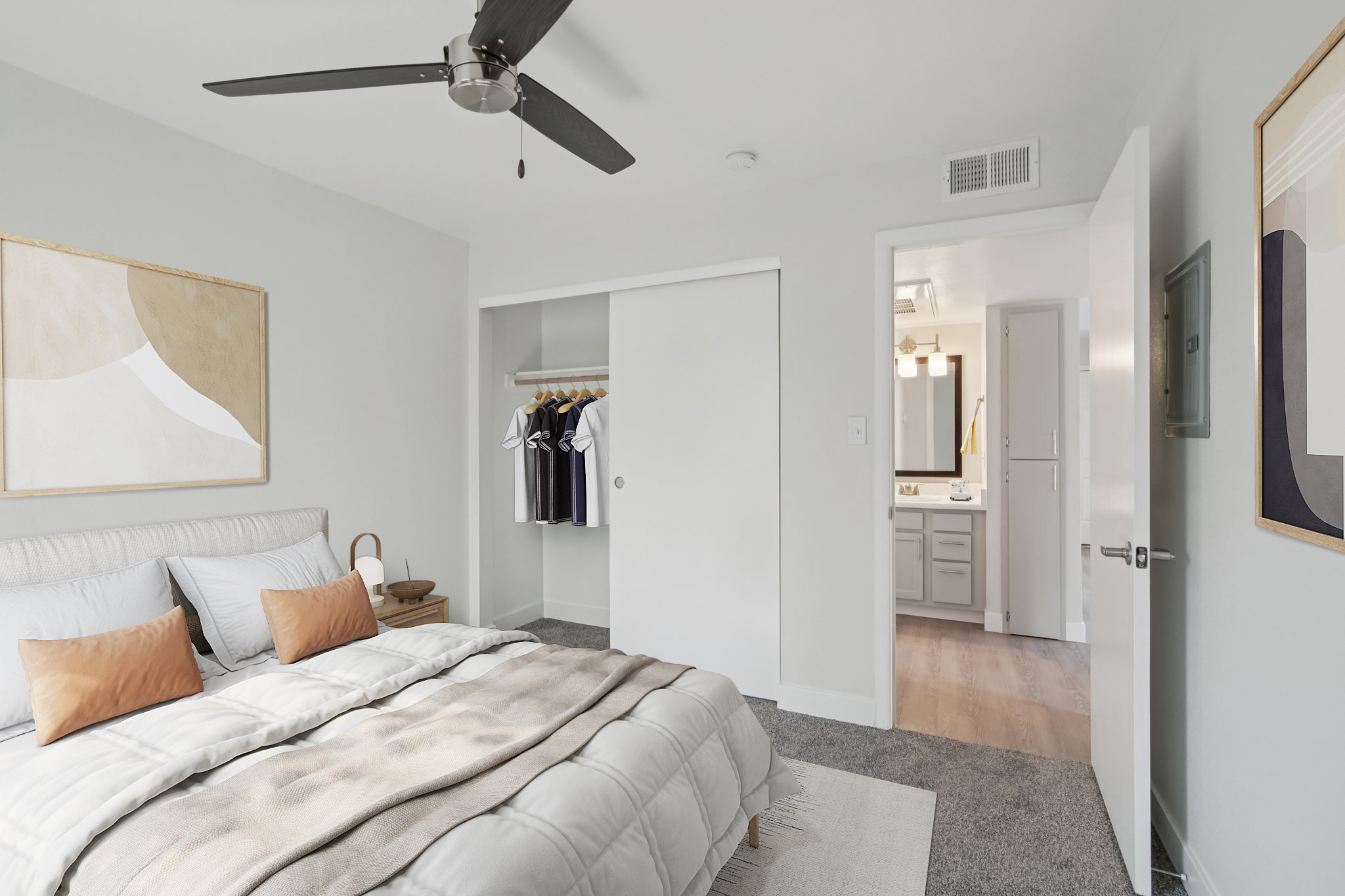 A modern bedroom featuring a neatly made bed with white and beige linens, two decorative orange pillows, a ceiling fan, and a closet with clothes on hangers. A bathroom is visible through an open door. The walls are light gray, and there are two framed artworks on display. The floor is carpeted.