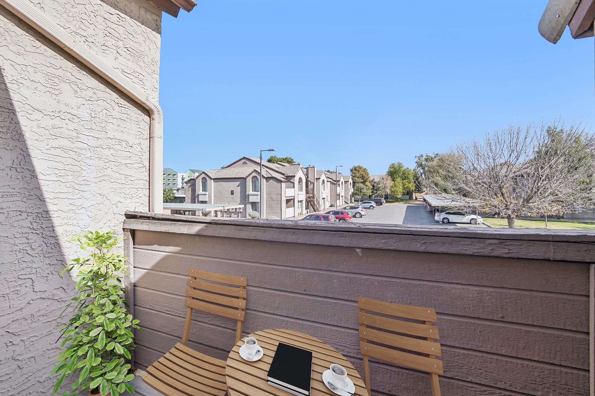 A cozy balcony view with a small wooden table and two chairs, featuring a coffee cup and a notebook. In the background, a row of residential buildings and parked cars under a clear blue sky. Foliage adds a touch of greenery to the scene.