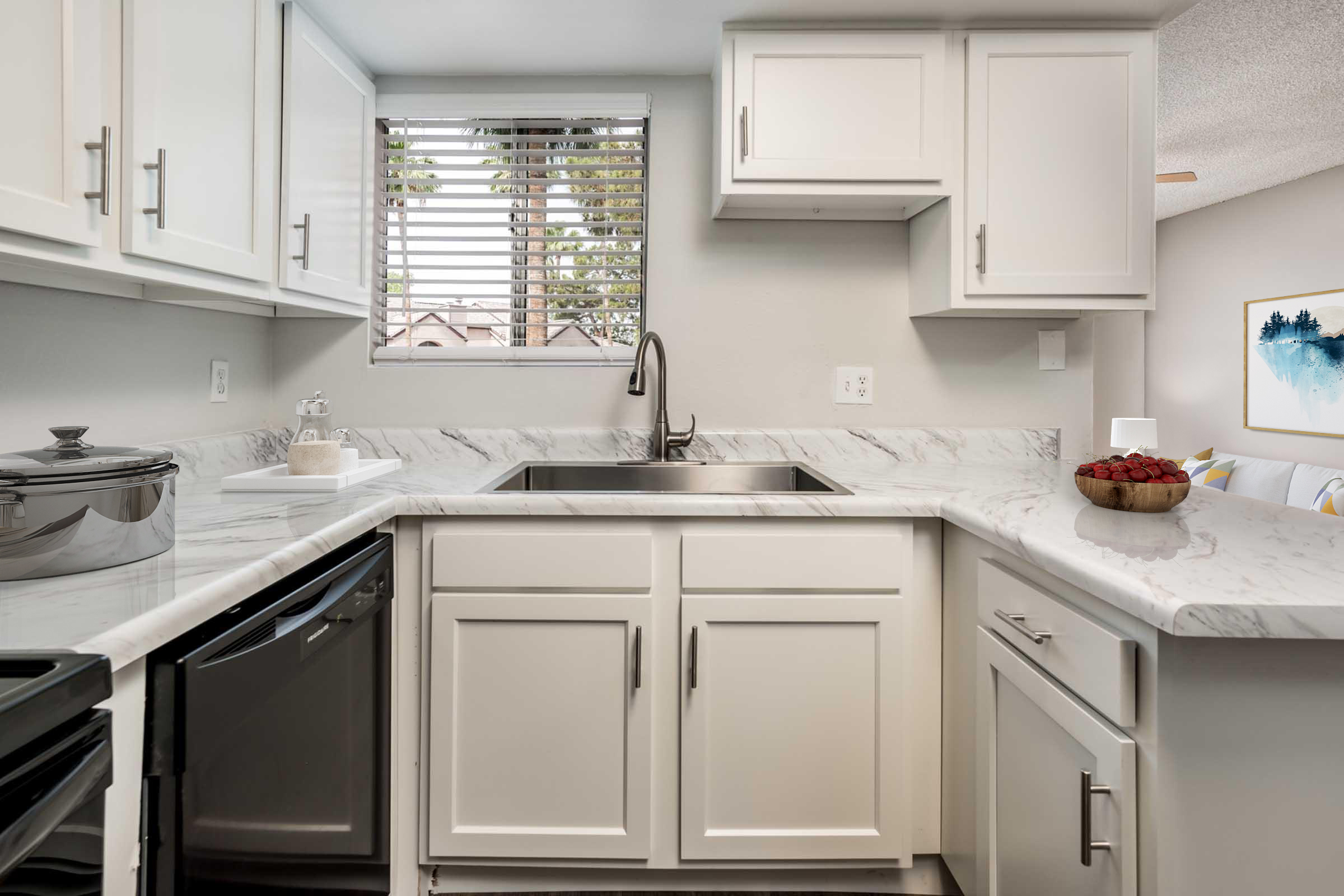 Modern kitchen featuring white cabinetry, a stainless steel sink, and a marble countertop. Natural light enters through a window with blinds. The kitchen includes a dishwasher and a countertop bowl filled with red fruits. A cozy living area is visible in the background with soft, neutral tones.