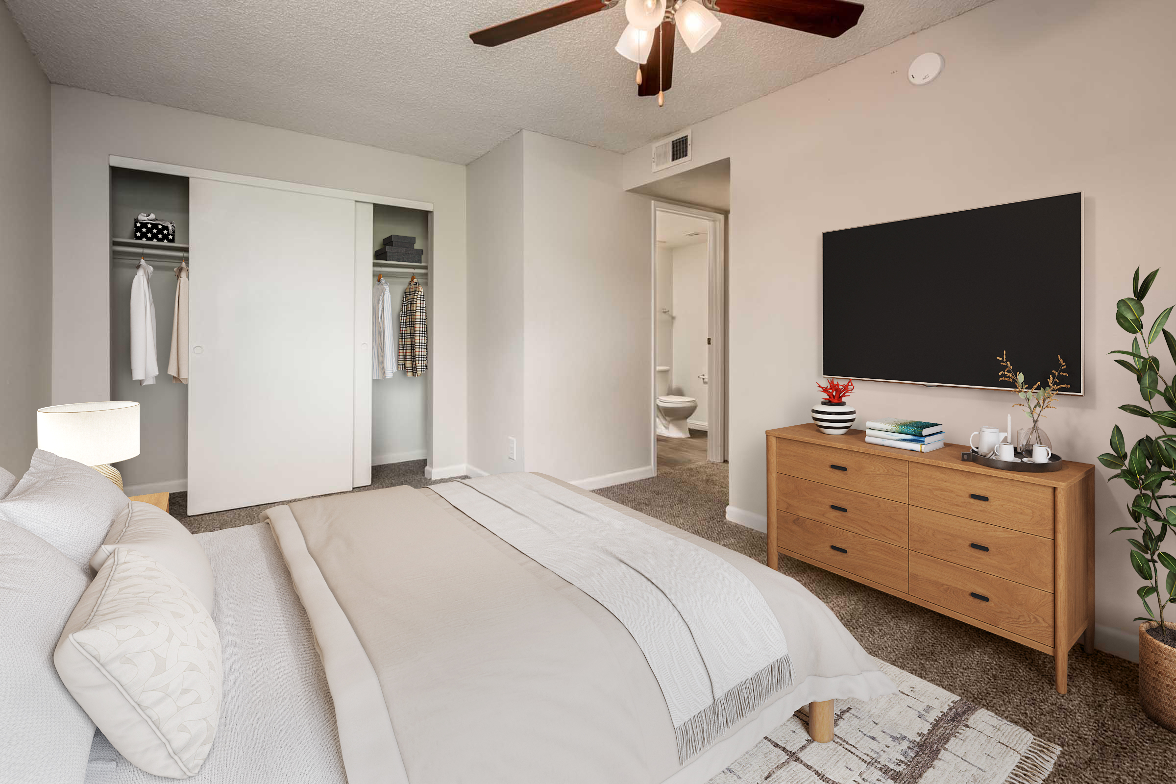 Cozy bedroom featuring a queen-sized bed with a light-colored blanket, a wooden dresser with decorative items, and a flat-screen TV mounted on the wall. A closet with sliding doors is visible, along with a doorway leading to a bathroom. The room is illuminated by a ceiling fan and has neutral-colored walls and carpet.