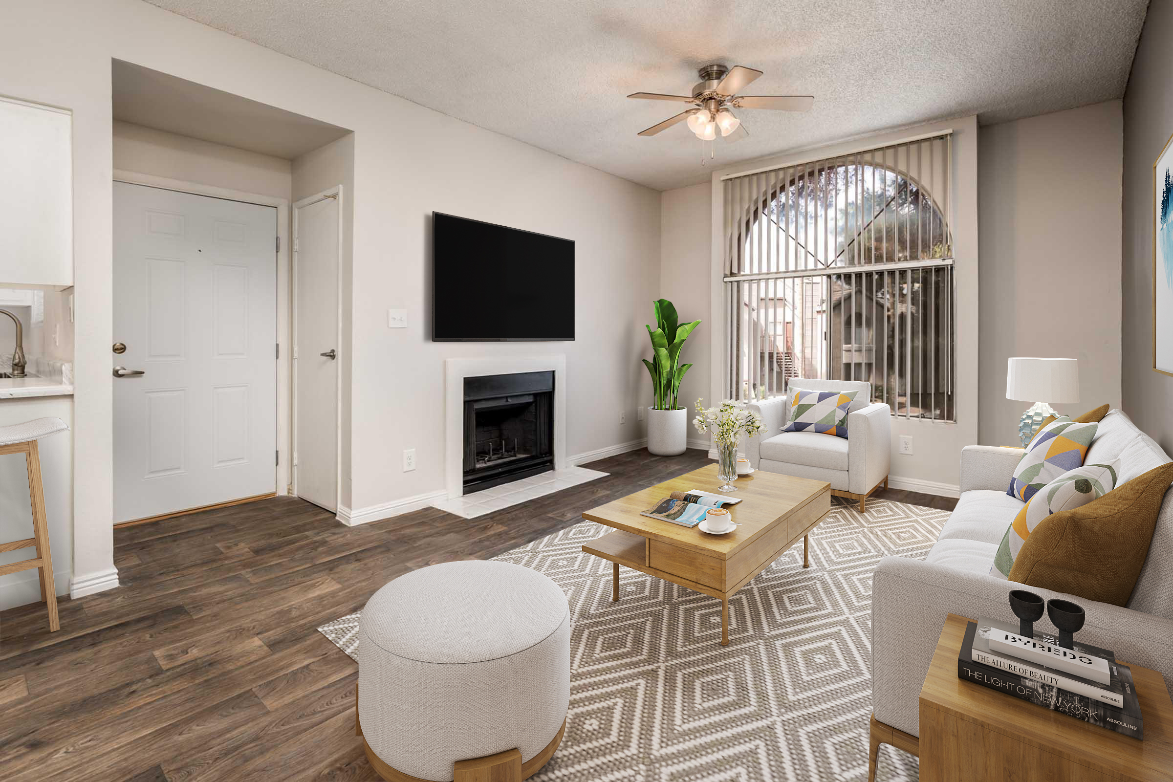 A bright and modern living room featuring a comfortable white sofa, a coffee table with decorative items, and a light-colored ottoman. A TV is mounted on the wall above a fireplace, and large windows with blinds allow natural light to fill the space. A green indoor plant adds a touch of nature to the room.