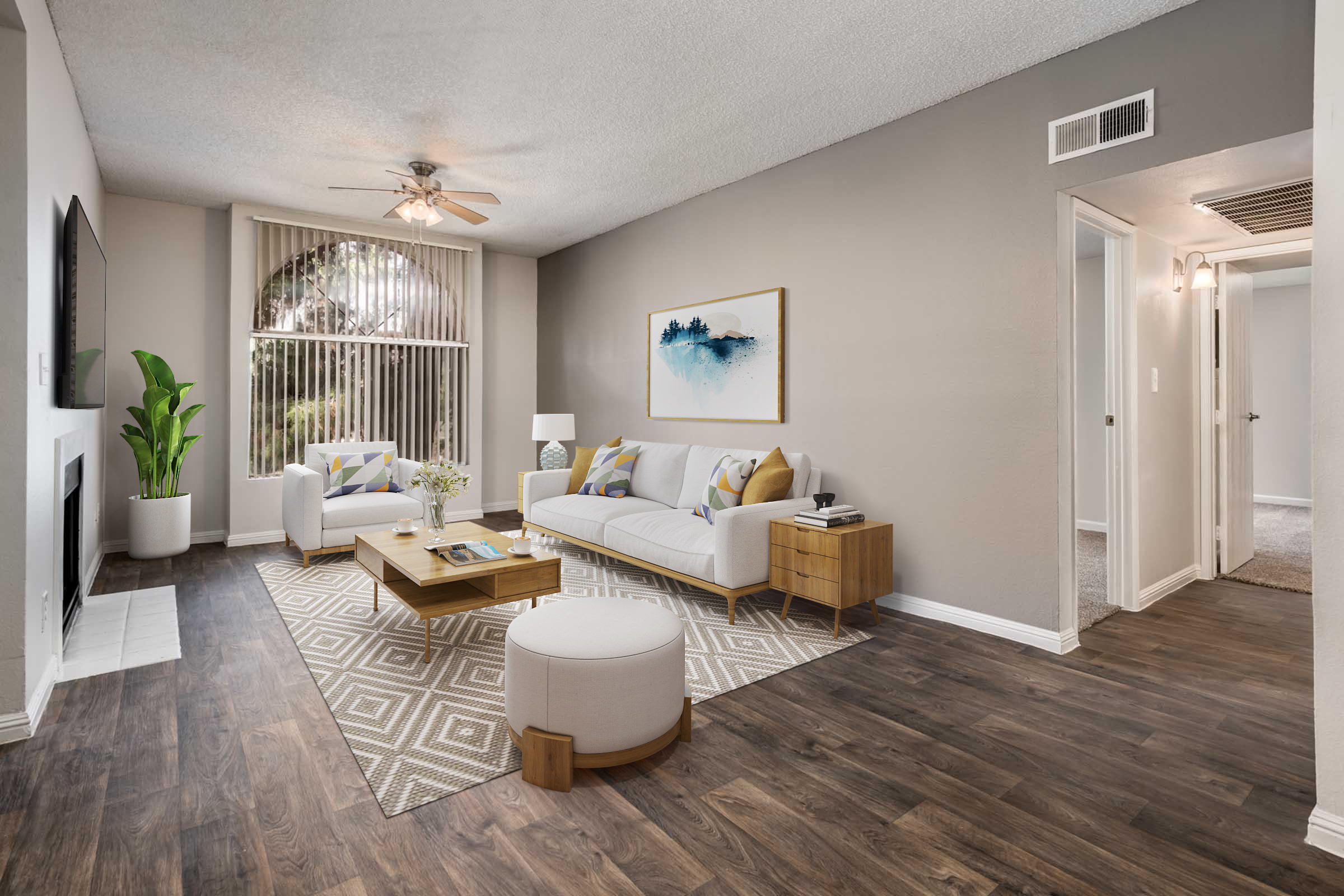 A modern living room featuring a white sofa with colorful throw pillows, a wooden coffee table, and a round ottoman. There is a plant in the corner, a flat-screen TV mounted on the wall, and large windows with blinds allowing natural light. The flooring is hardwood, and the walls are painted a soft gray.