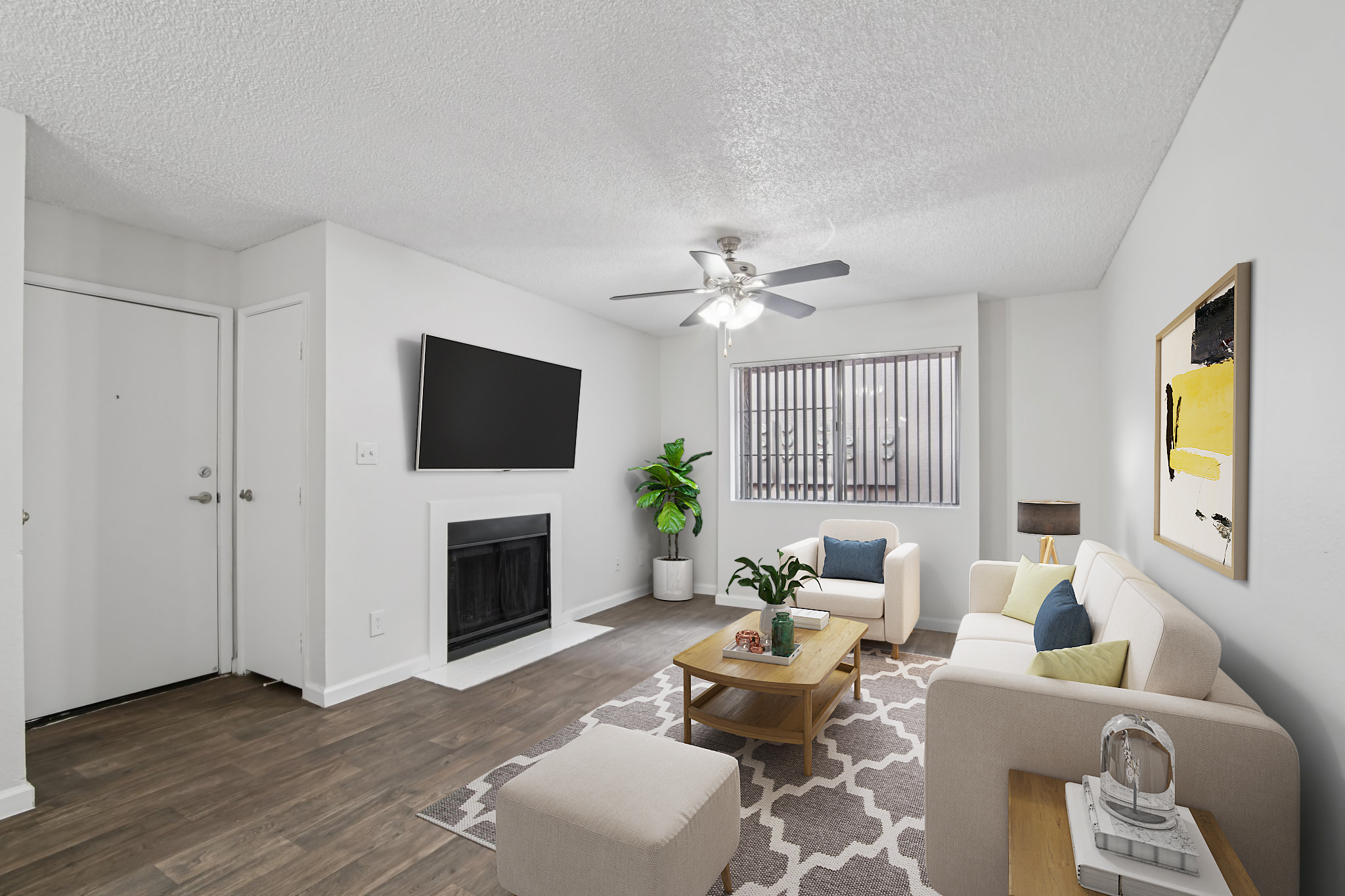 A modern, well-lit living room featuring a beige sofa, a coffee table, and an ottoman. There's a flat-screen TV mounted on the wall, a potted plant in the corner, and a fireplace beneath a window with vertical blinds. The room has neutral walls and wooden flooring, creating a cozy atmosphere.