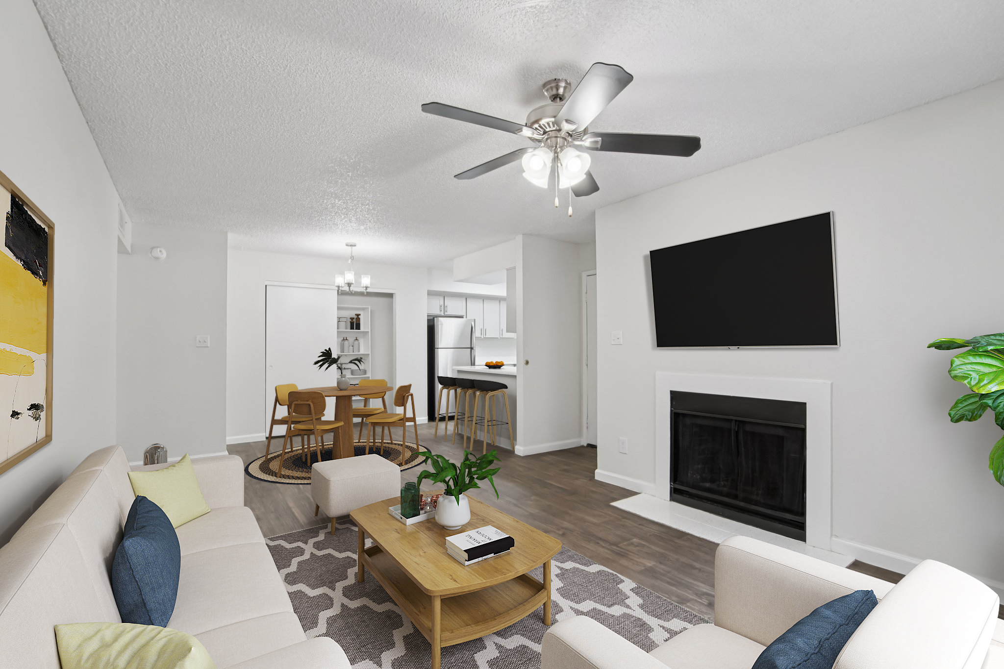 A modern living room featuring a light gray couch with colorful throw pillows, a wooden coffee table, and a ceiling fan. The room includes a mounted flat-screen TV, a cozy fireplace, and an open view into a dining area with a round table and chairs. Natural light brightens the space.