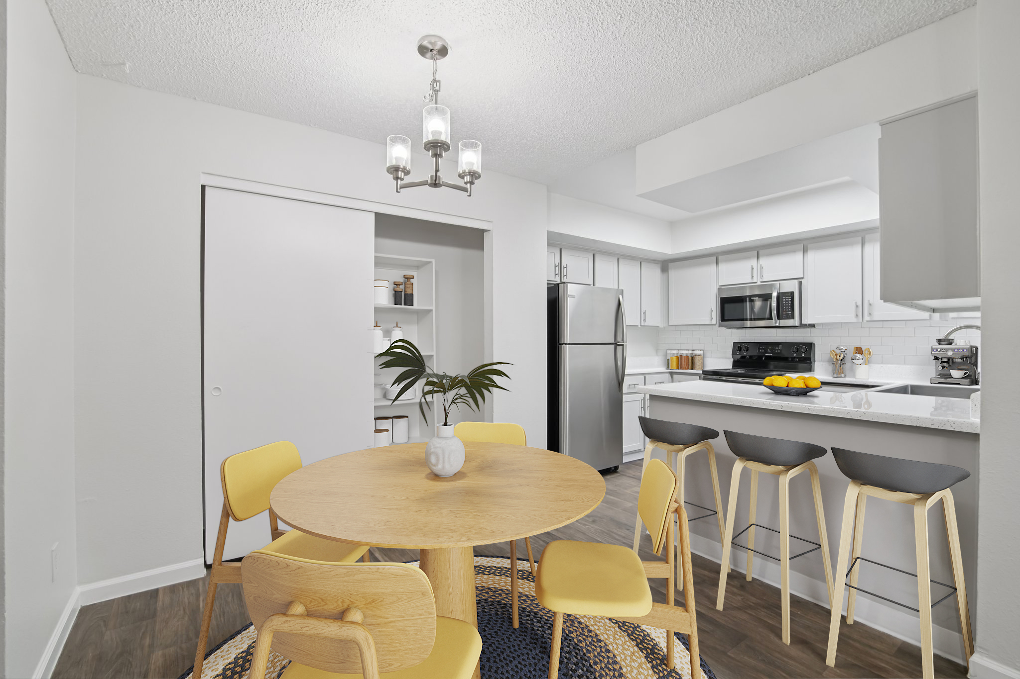 A modern kitchen and dining area featuring a round wooden table with yellow chairs, a rug underneath, a small plant as a centerpiece, and a bar counter with stools. The kitchen is bright and has stainless steel appliances, cabinets, and a window, creating a welcoming and functional space.
