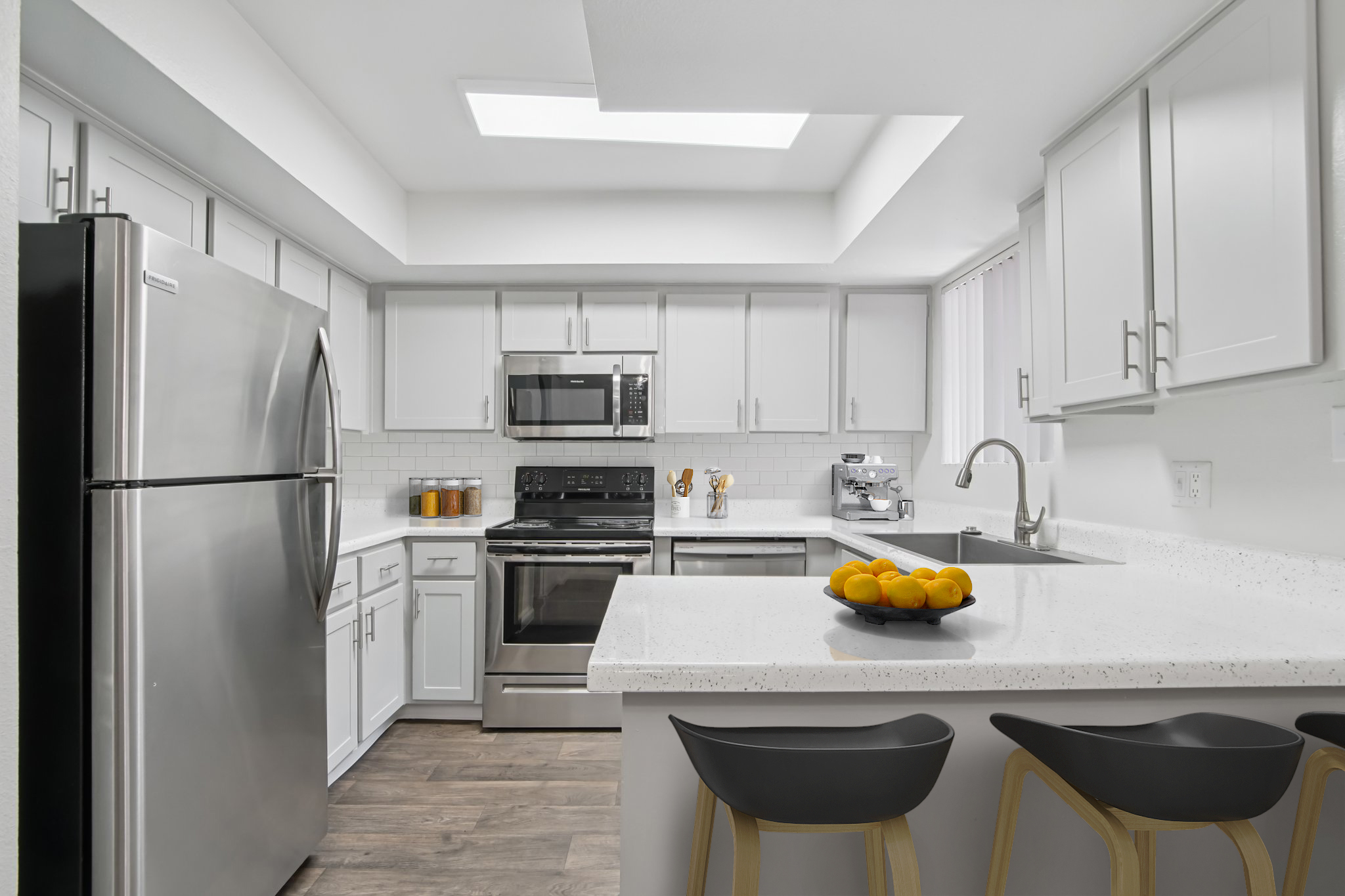 Modern kitchen featuring stainless steel appliances, including a refrigerator and oven. The white cabinetry contrasts with the dark countertops, and a bowl of oranges is placed on the island. A cozy dining area is visible, complemented by natural light streaming in through a skylight.