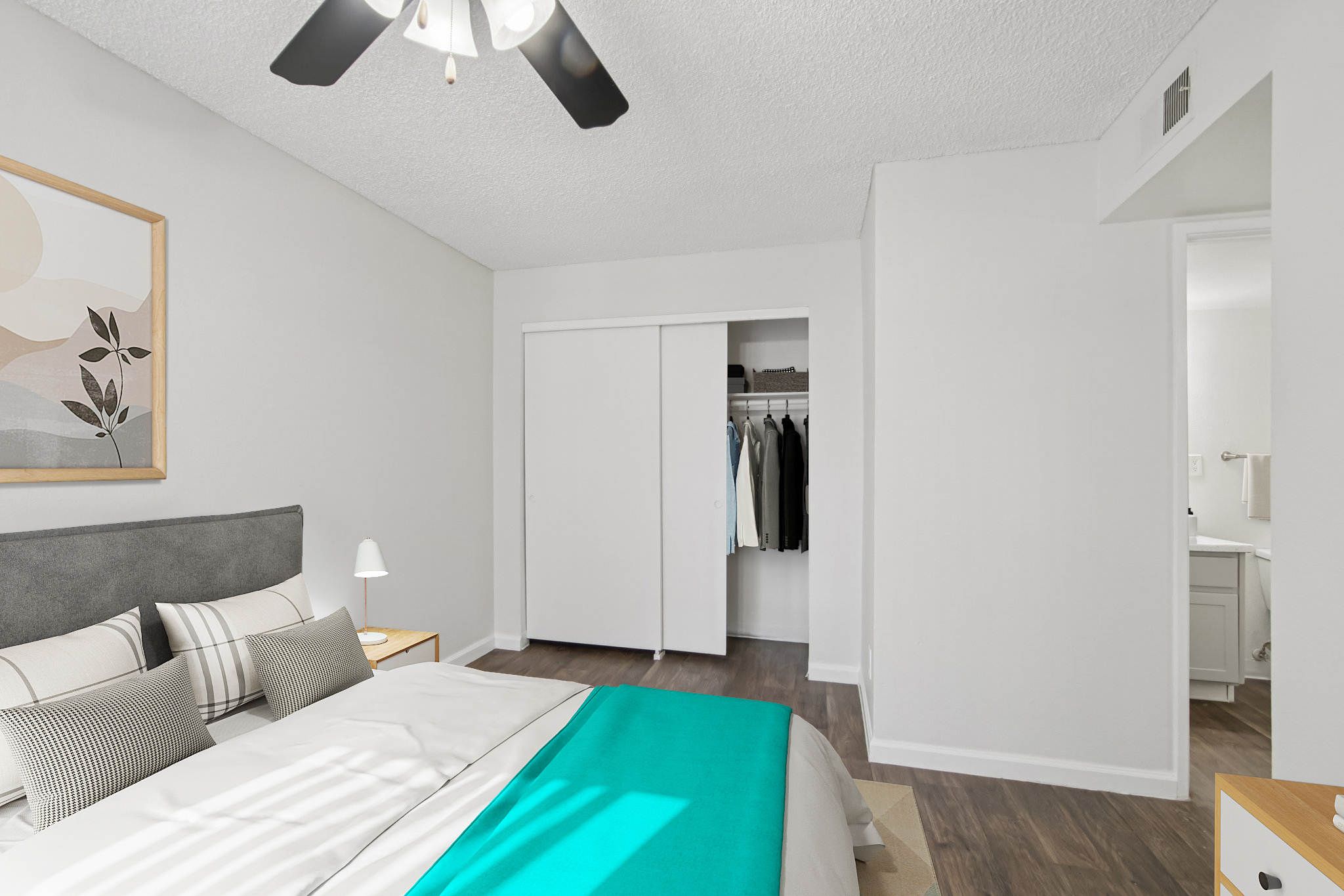 A modern bedroom featuring a large bed with a teal throw blanket, bedside lamp, and framed artwork on the wall. A closet with white sliding doors is visible, showcasing neatly hung clothes. The room has light-colored walls and wood flooring, creating a bright and inviting atmosphere.