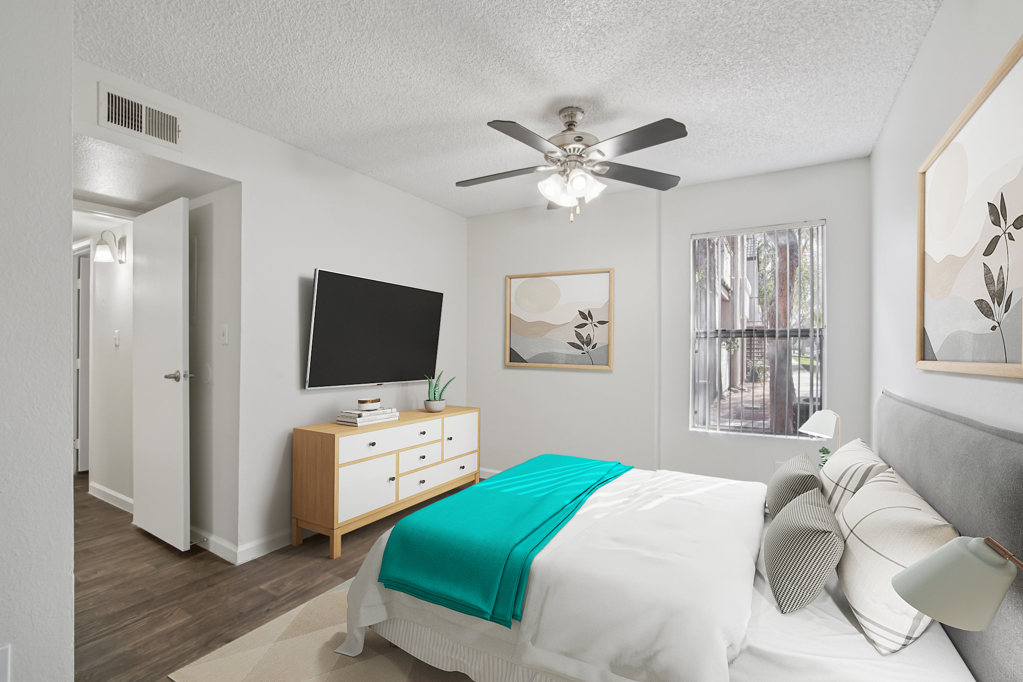 A cozy bedroom featuring a bed with a light-colored duvet and a turquoise throw. There is a ceiling fan above, a wall-mounted TV, and a wooden dresser. Decor includes framed artwork and a window with natural light. The room has light-colored walls and modern furnishings, creating a welcoming atmosphere.