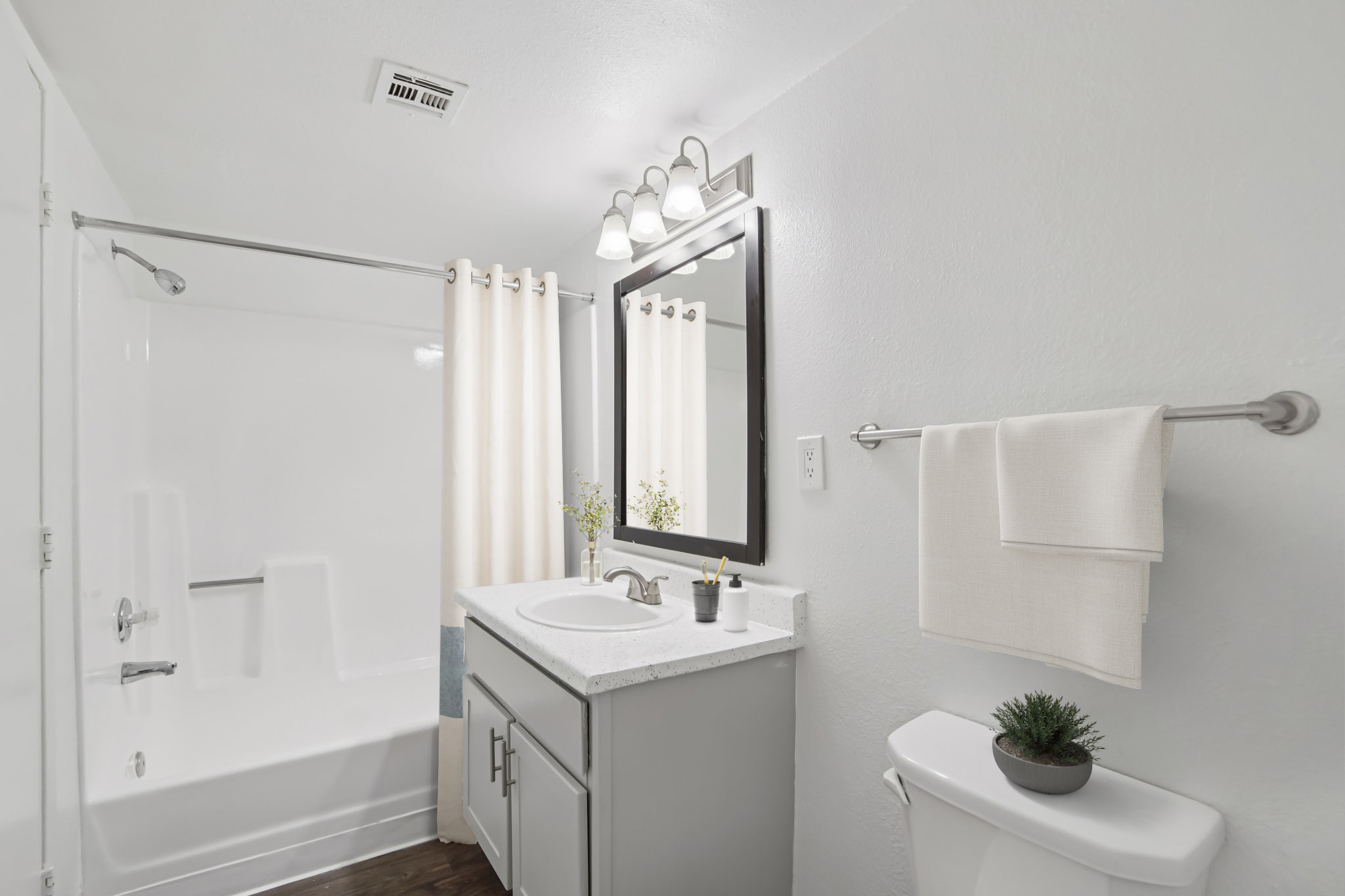 A modern bathroom featuring a bathtub with a shower, a white countertop with sink, a large mirror, and decorative touches. The walls are painted white, and there is a towel rack with a neatly hung towel and a small plant on the toilet tank. Natural light and a clean design create a bright atmosphere.