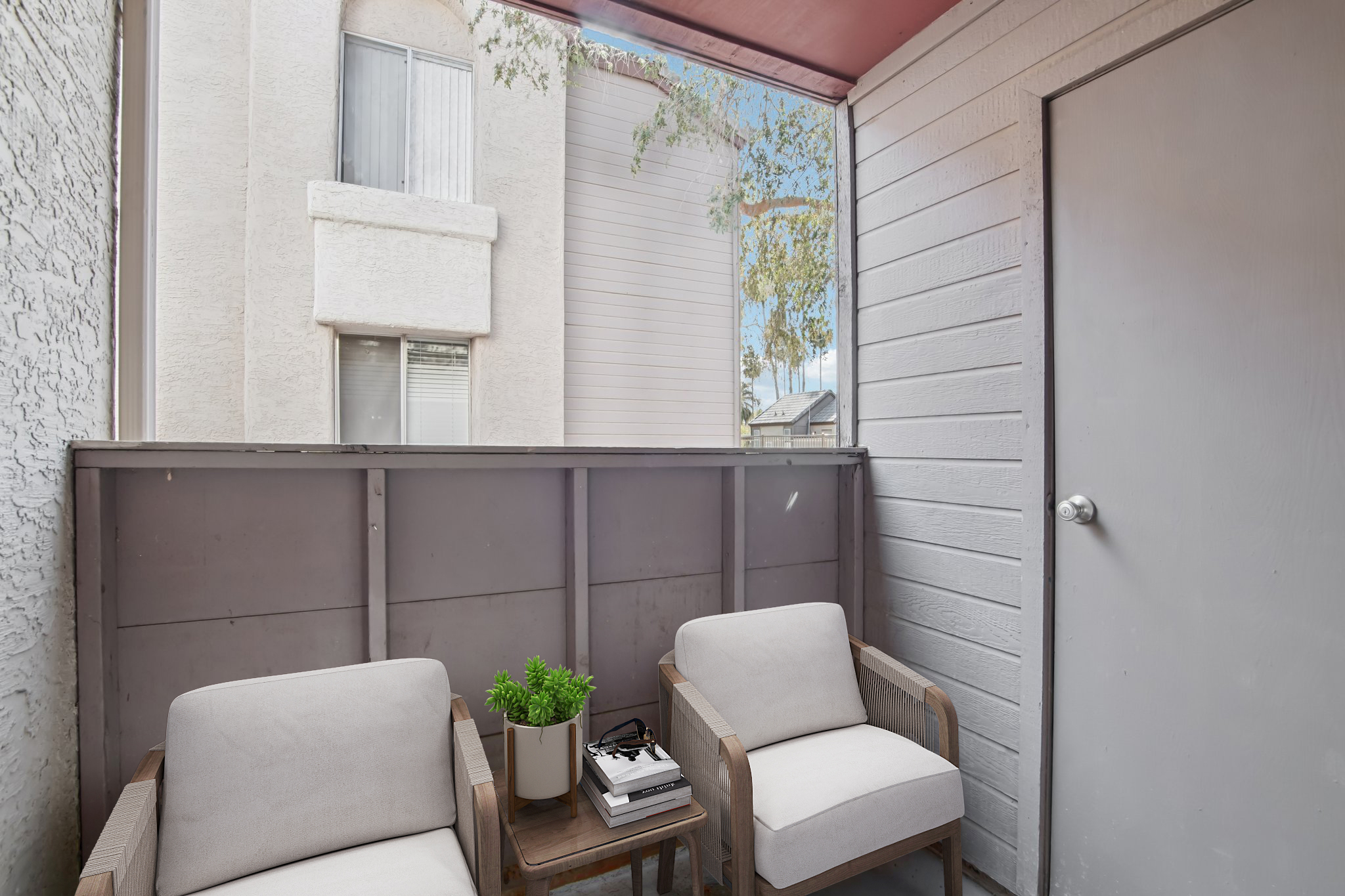 A small balcony featuring two cozy armchairs with light cushions, a table with a plant and a book, and a view of adjacent building walls. The balcony has a wooden railing and a partially covered ceiling, creating a private and inviting atmosphere.