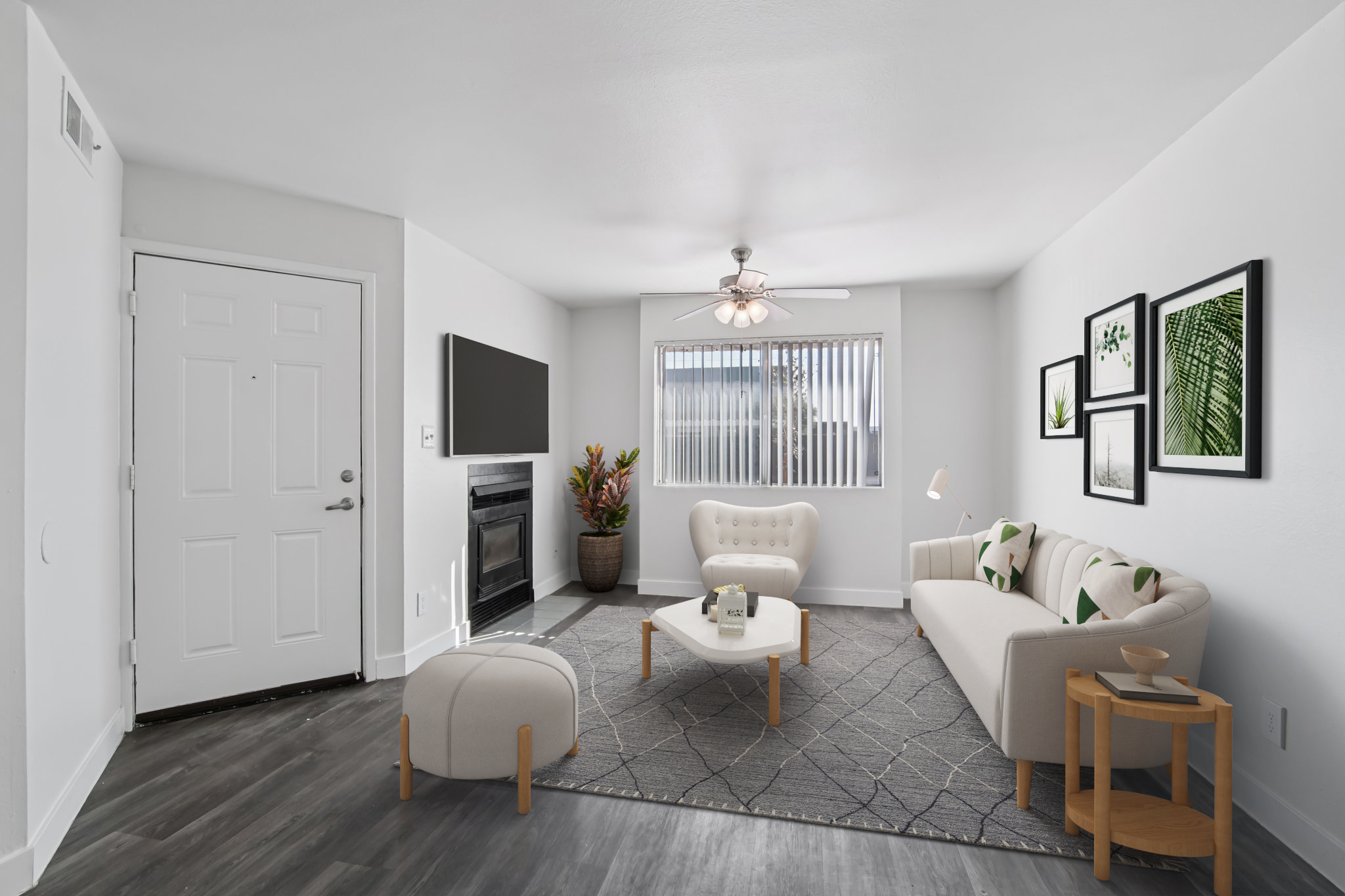 A bright and modern living room featuring a light-colored sofa and armchair, a coffee table, and a TV mounted on the wall. Large windows allow natural light to enter, with decorative plants and framed artwork on the walls. The flooring is a warm gray tone, adding to the inviting atmosphere.