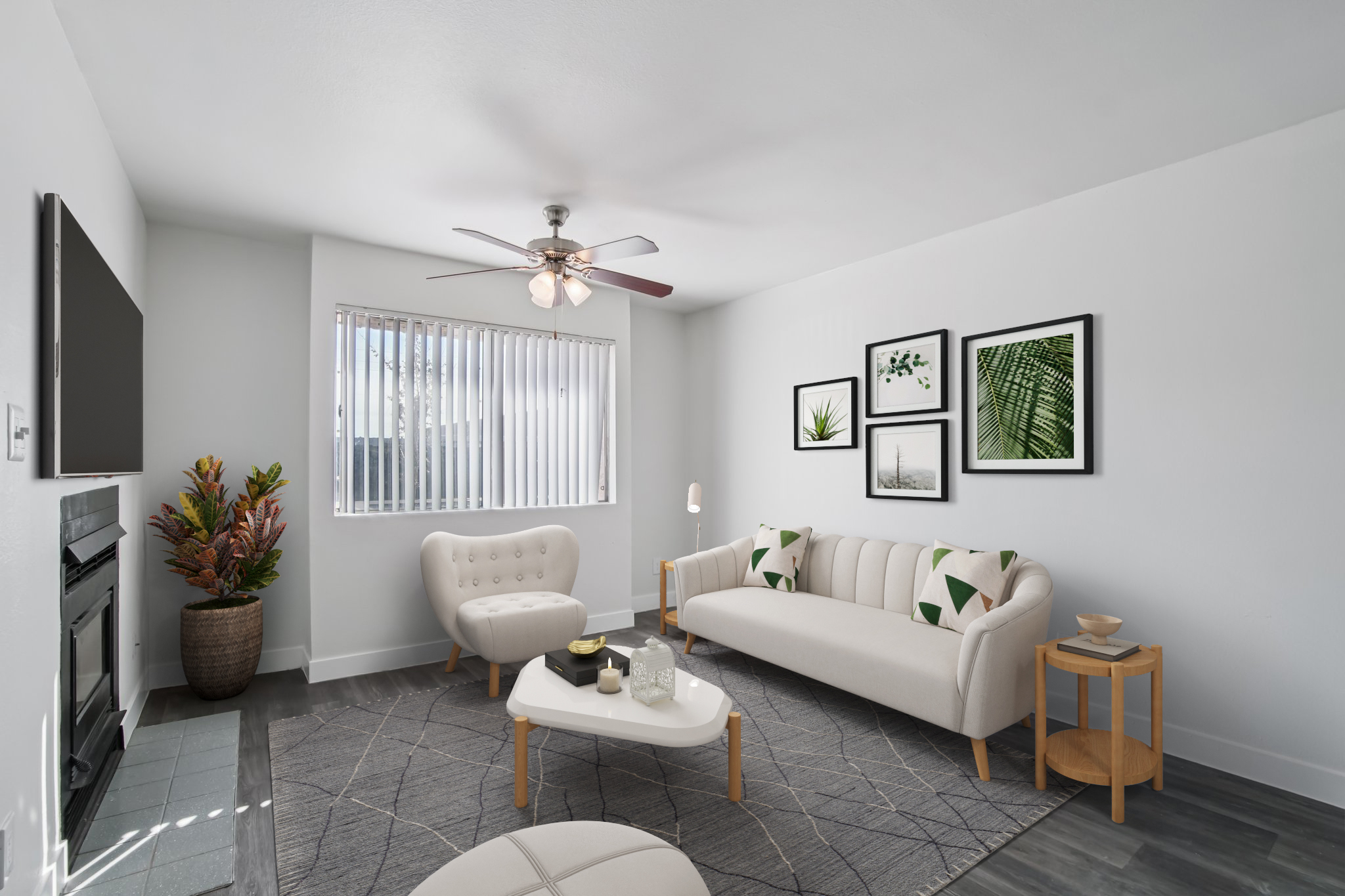A modern living room with light-colored furniture including a sofa and armchair. The space features a ceiling fan, a flat-screen TV on the wall, and decorative plants. Large windows with blinds allow natural light, complemented by framed botanical artwork on the walls. The floor is covered with a geometric patterned rug.