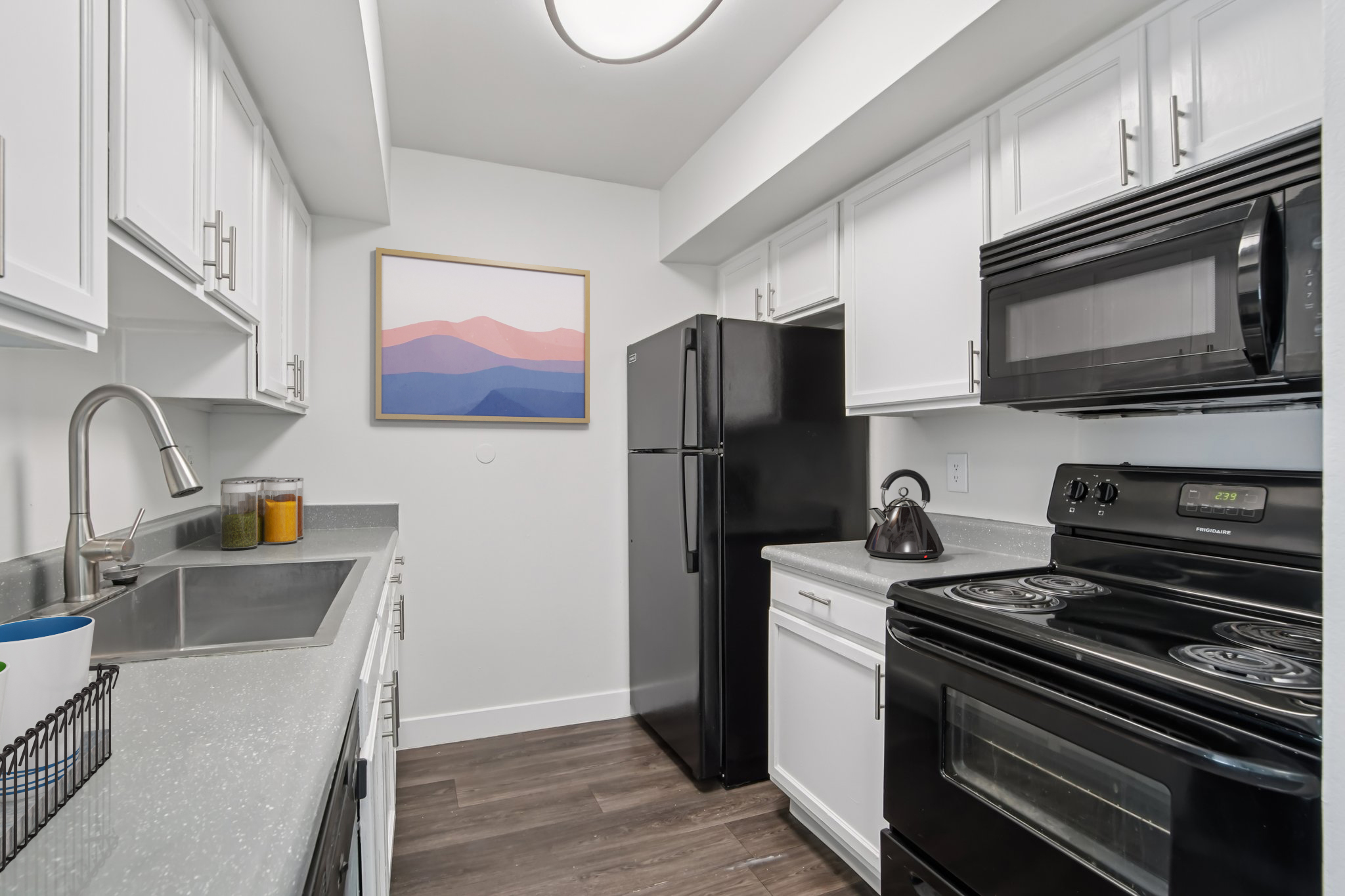 A modern kitchen featuring white cabinetry, a black refrigerator, and a black stove. The countertops are light gray with a sink and a decorative painting of mountains on the wall above. The flooring is a dark wood look, and there are some colorful jars on the counter. Well-lit and neatly organized space.