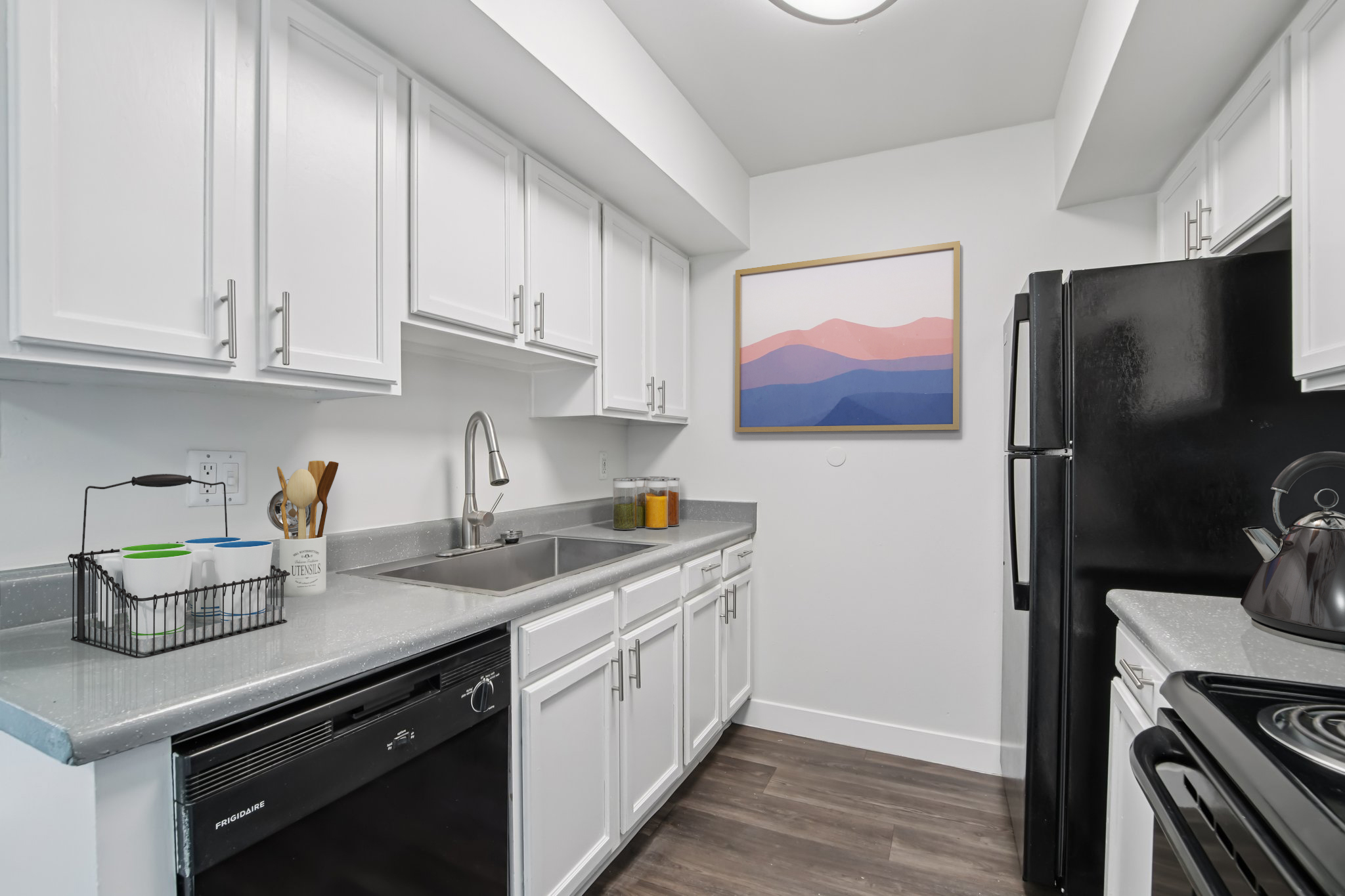 Modern kitchen featuring white cabinets, a black refrigerator, and a sink with stainless steel fixtures. The countertop is gray, and there's a black dishwasher underneath. A framed art piece with soft pink and blue hues hangs on the wall, adding a touch of color to the space. Wood-like flooring completes the contemporary look.