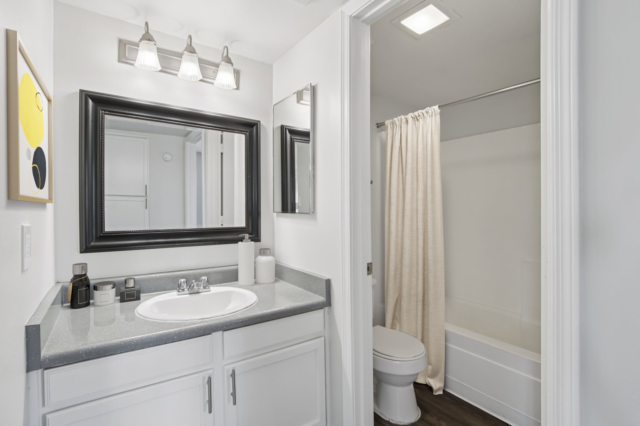 A modern bathroom featuring a double sink vanity with a gray countertop, wall-mounted mirrors, and stylish lighting. A shower/tub combo is seen behind a cream-colored curtain, while the space is bright and well-organized with decorative items on the counter.