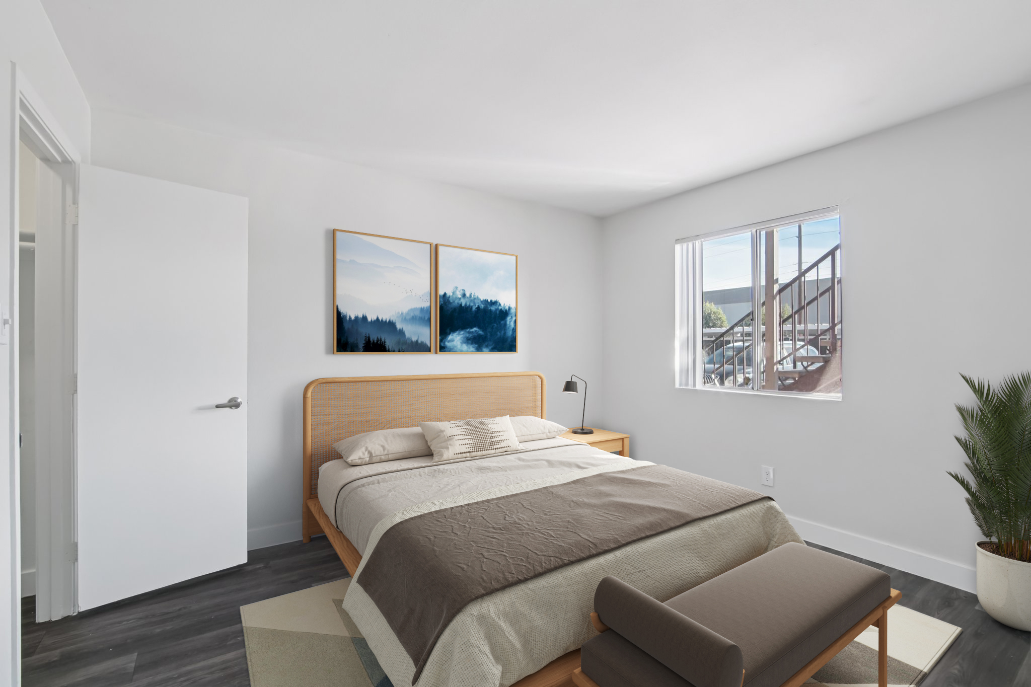 A modern, minimalistic bedroom featuring a wooden bed with neutral bedding, a nightstand with a lamp, and two framed landscape images on the wall. There’s a window allowing natural light, a small plant near the bed, and a light gray rug on the floor, creating a cozy and inviting atmosphere.