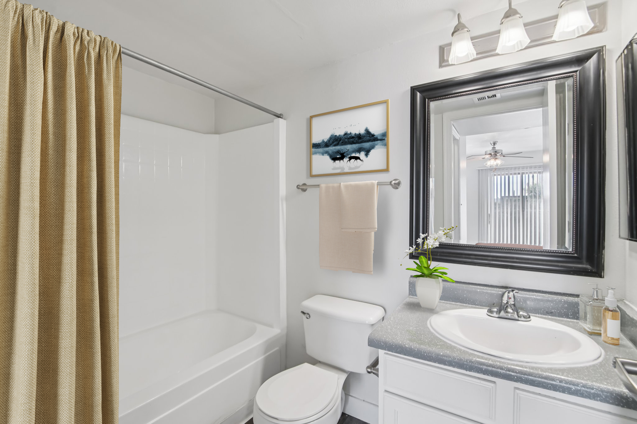 Modern bathroom featuring a bathtub with a shower curtain, a white toilet, and a stylish vanity with a sink. The walls are painted white, and a framed picture hangs above the sink. A mirror with a black frame reflects the space, complemented by a small plant and decorative items on the countertop.