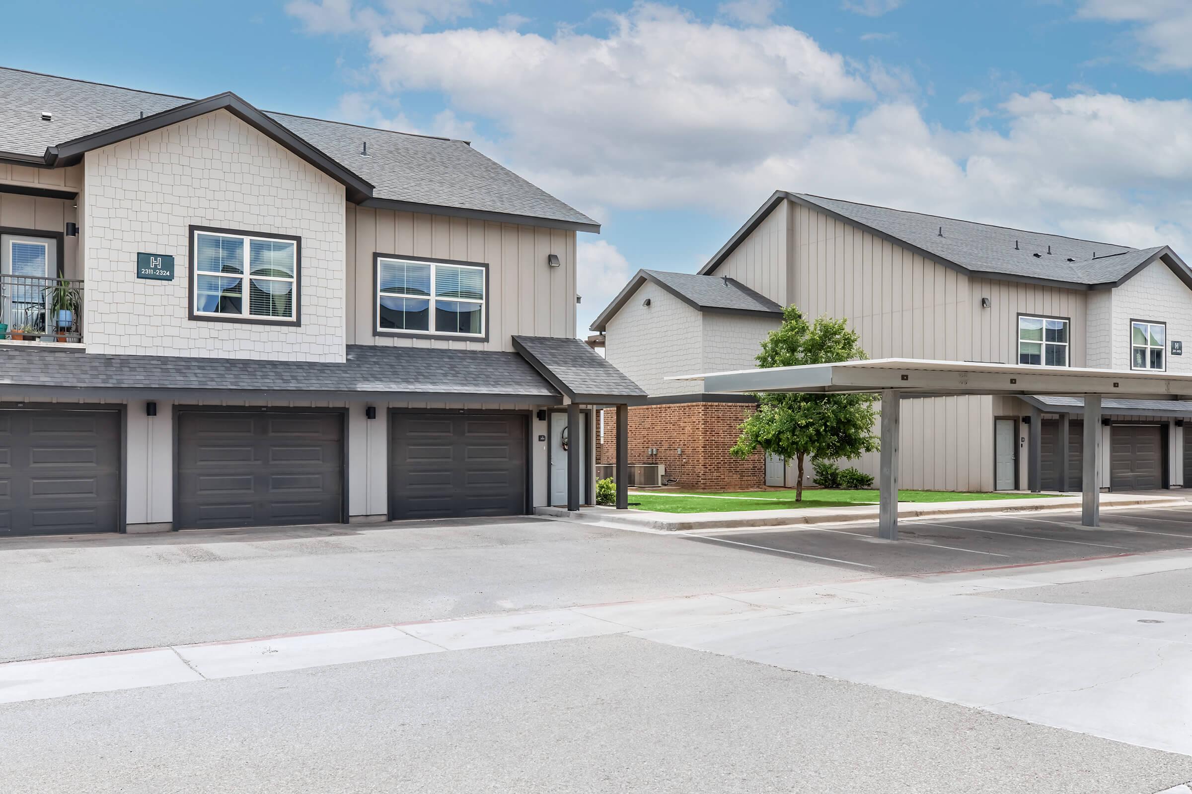 A residential neighborhood featuring modern two-story buildings with gray exteriors, featuring garages. One building has balconies, while another showcases a carport area with green lawns and trees nearby. The sky is partly cloudy, enhancing the bright and inviting atmosphere.