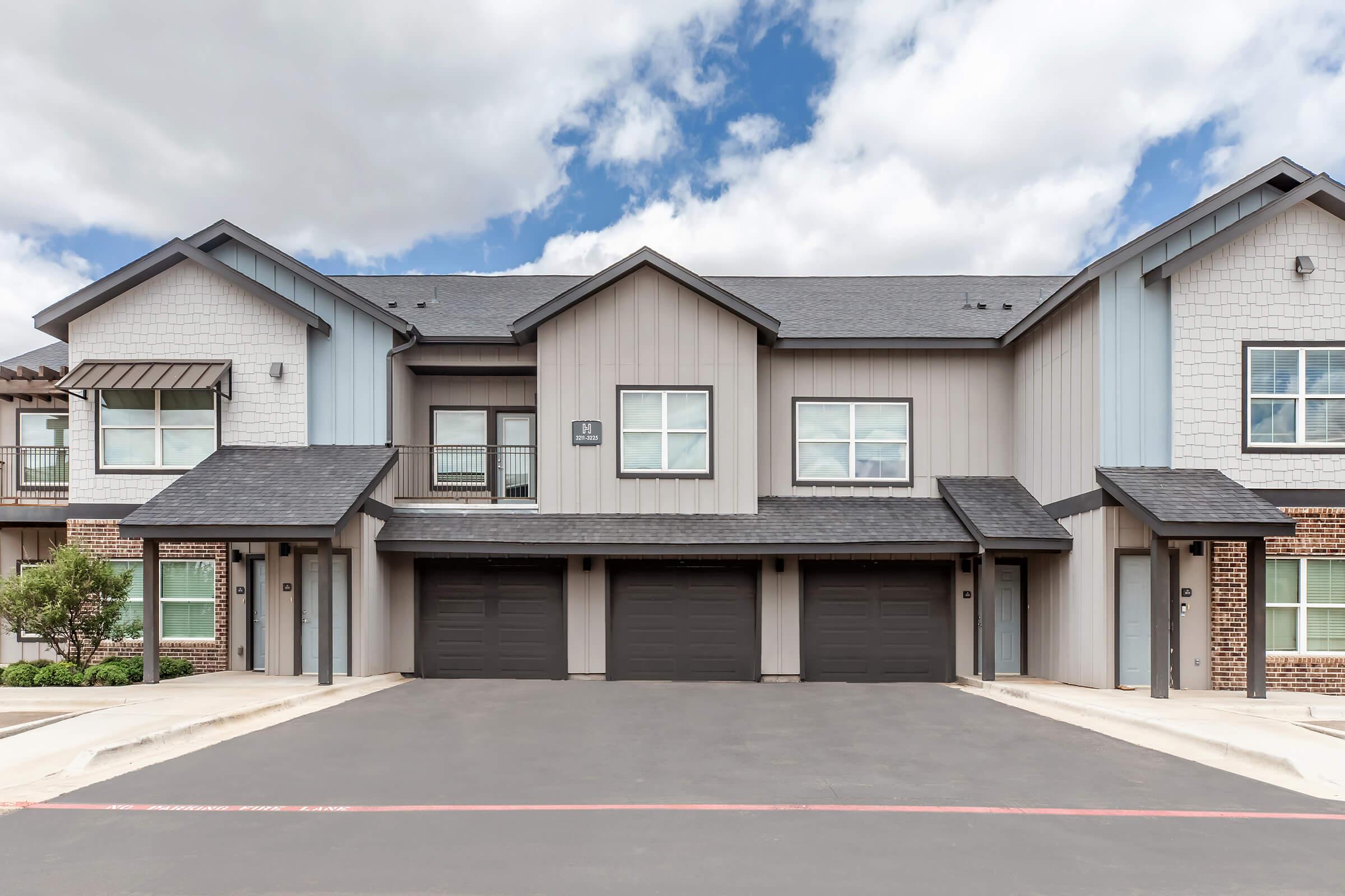 Modern two-story apartment complex with a combination of gray and beige siding. The building features multiple units with large windows, black garage doors, and a well-maintained parking area. The sky is partly cloudy, adding a pleasant backdrop to the scene.