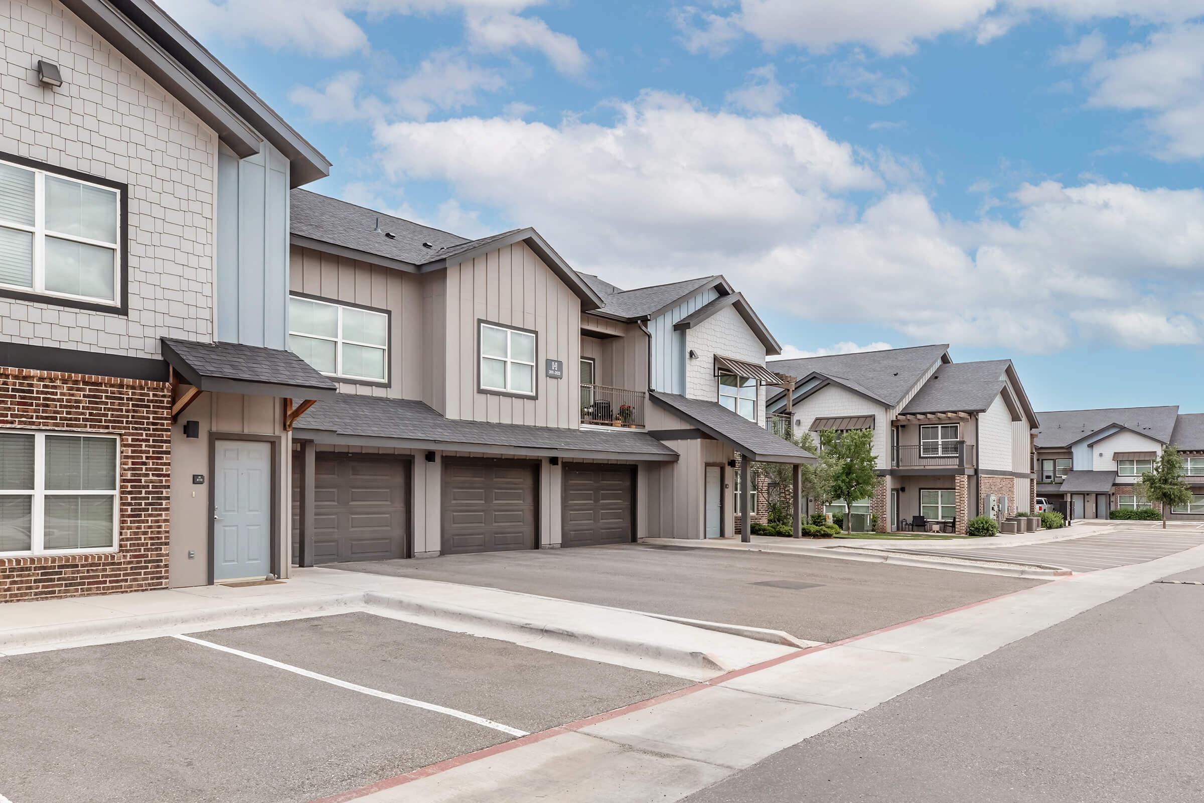 A modern residential neighborhood featuring two-story townhouses with a blend of brick and siding exteriors. The units have garages and are arranged along a paved street with landscaped patches. The sky is partly cloudy, adding a pleasant ambiance to the setting.