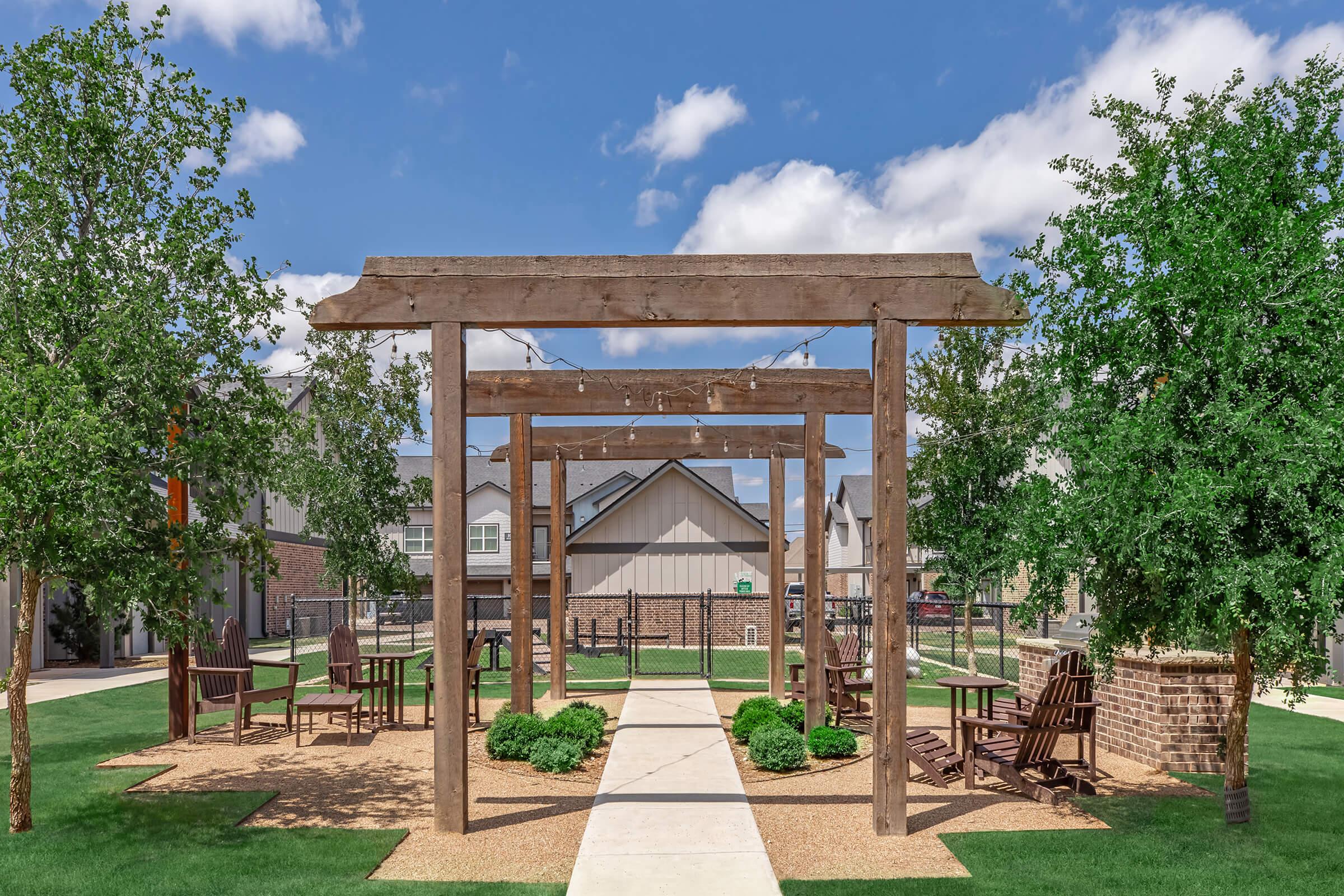 A well-maintained outdoor seating area featuring wooden pergolas, surrounded by lush green shrubs and trees. A paved walkway leads through the space, with several wooden chairs placed around small tables. The sky is clear with a few fluffy clouds, and residential buildings are visible in the background.