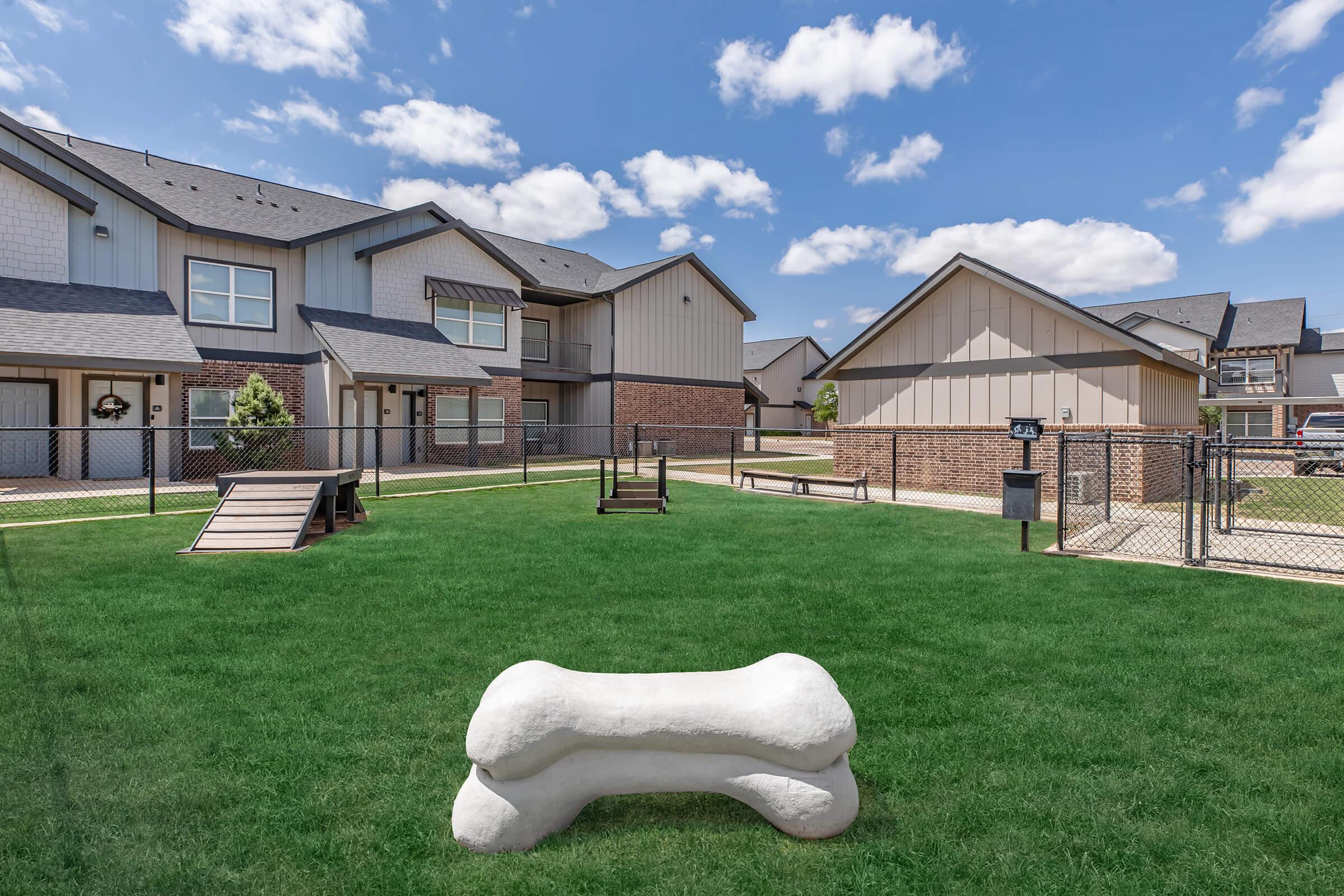 A spacious outdoor area featuring a grassy lawn with a large white dog bone sculpture, agility exercise equipment, and modern residential buildings in the background. The sky is clear with fluffy clouds, creating a bright and inviting environment for pets and their owners.