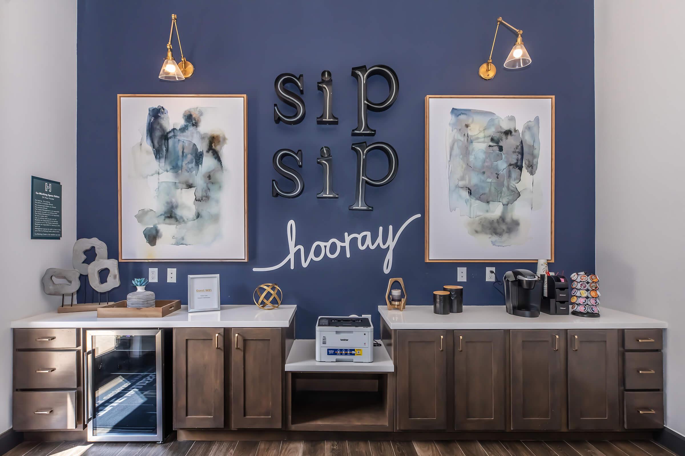A stylish coffee station featuring a dark blue wall with two abstract art pieces framed in gold. The words "sip sip hooray" are artistically displayed. Below, there are cabinets containing a coffee maker, cups, and an inviting atmosphere with modern light fixtures.