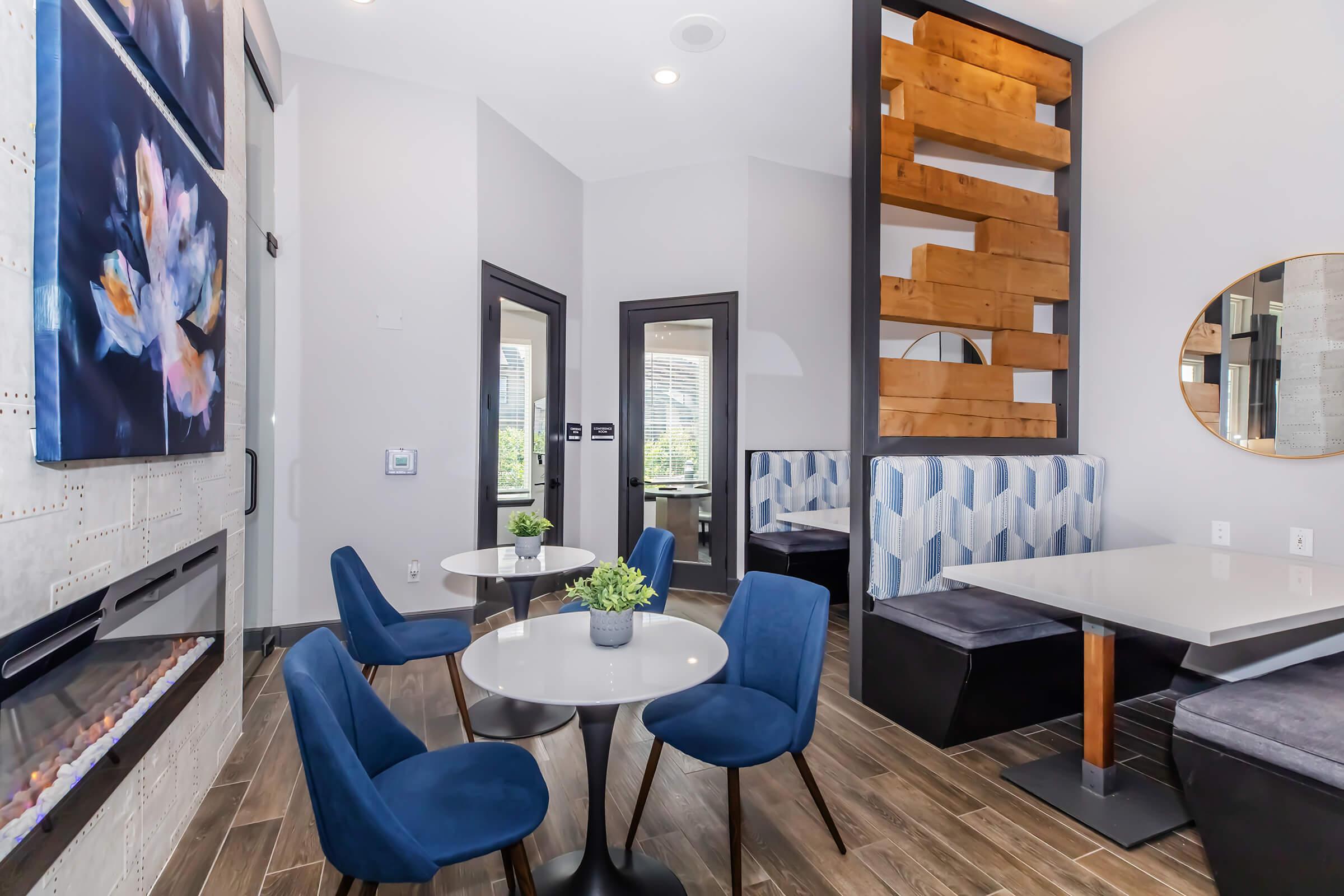 Modern cafe interior featuring a stylish design with blue chairs and tables, a cozy corner with wooden accents, and large windows allowing natural light. Decorative elements include a wall-mounted fireplace and vibrant artwork.