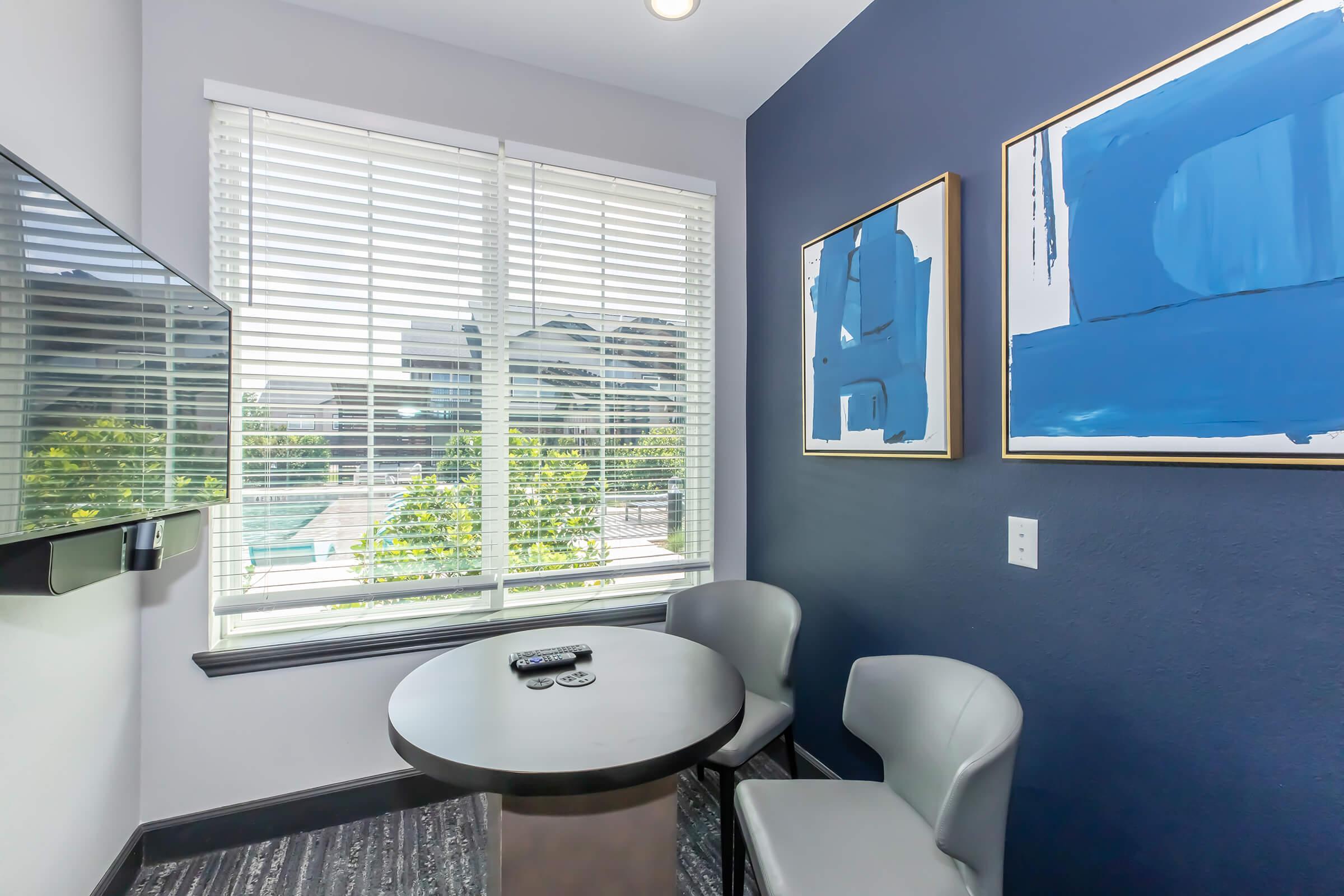A small room featuring a round table with two gray chairs, a mounted television on the wall, and large windows adorned with white blinds. The walls are painted dark blue with two abstract blue artworks framed and hung. Greenery can be seen outside the windows, adding a touch of nature to the minimalist space.
