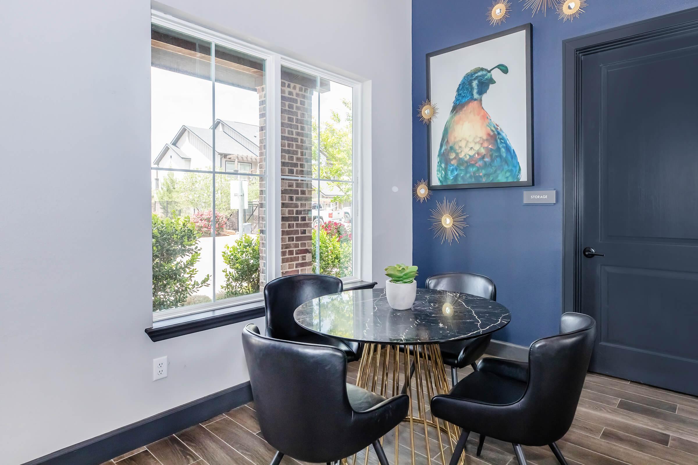 A stylish dining area featuring a round black marble table surrounded by four black chairs. A colorful bird painting hangs on the blue wall, and there are large windows providing natural light and a view of greenery outside. The floor is made of wood, creating a warm atmosphere.