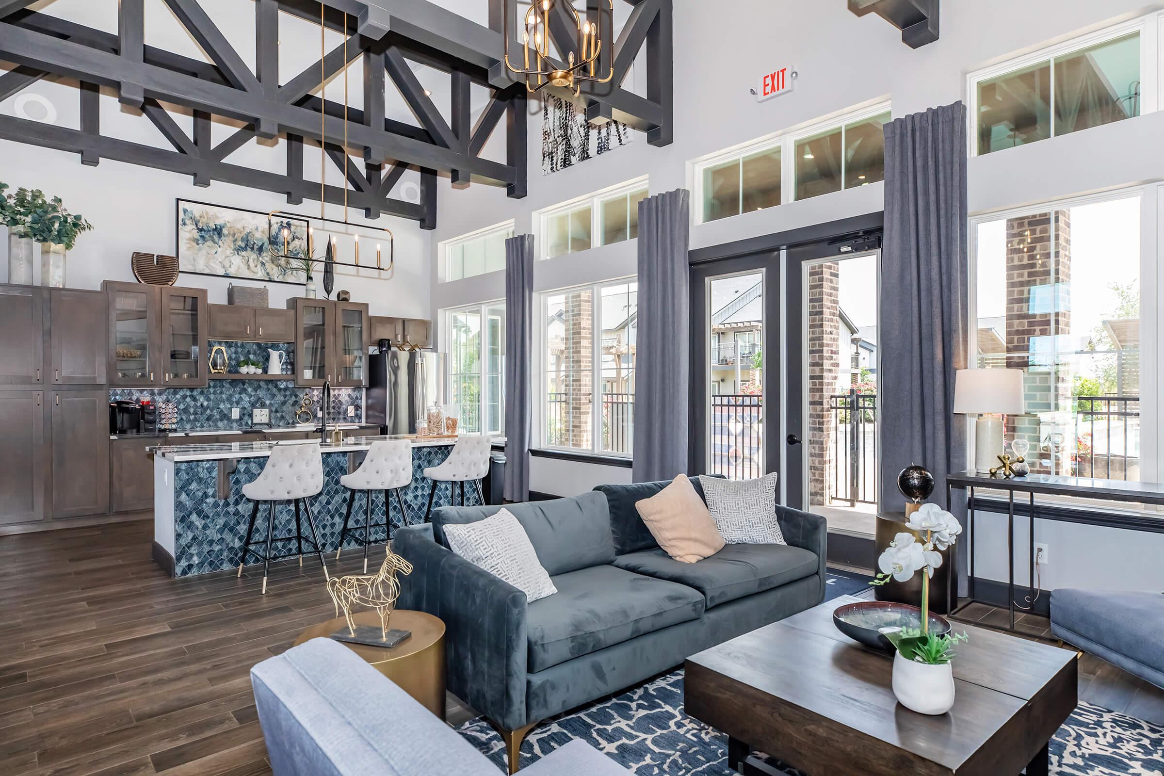 A modern living area featuring a stylish sofa with decorative pillows, a coffee table, and a large window displaying outdoor views. The kitchen area has bar stools and a blue mosaic backsplash. High ceilings with exposed beams and elegant light fixtures create a spacious atmosphere.