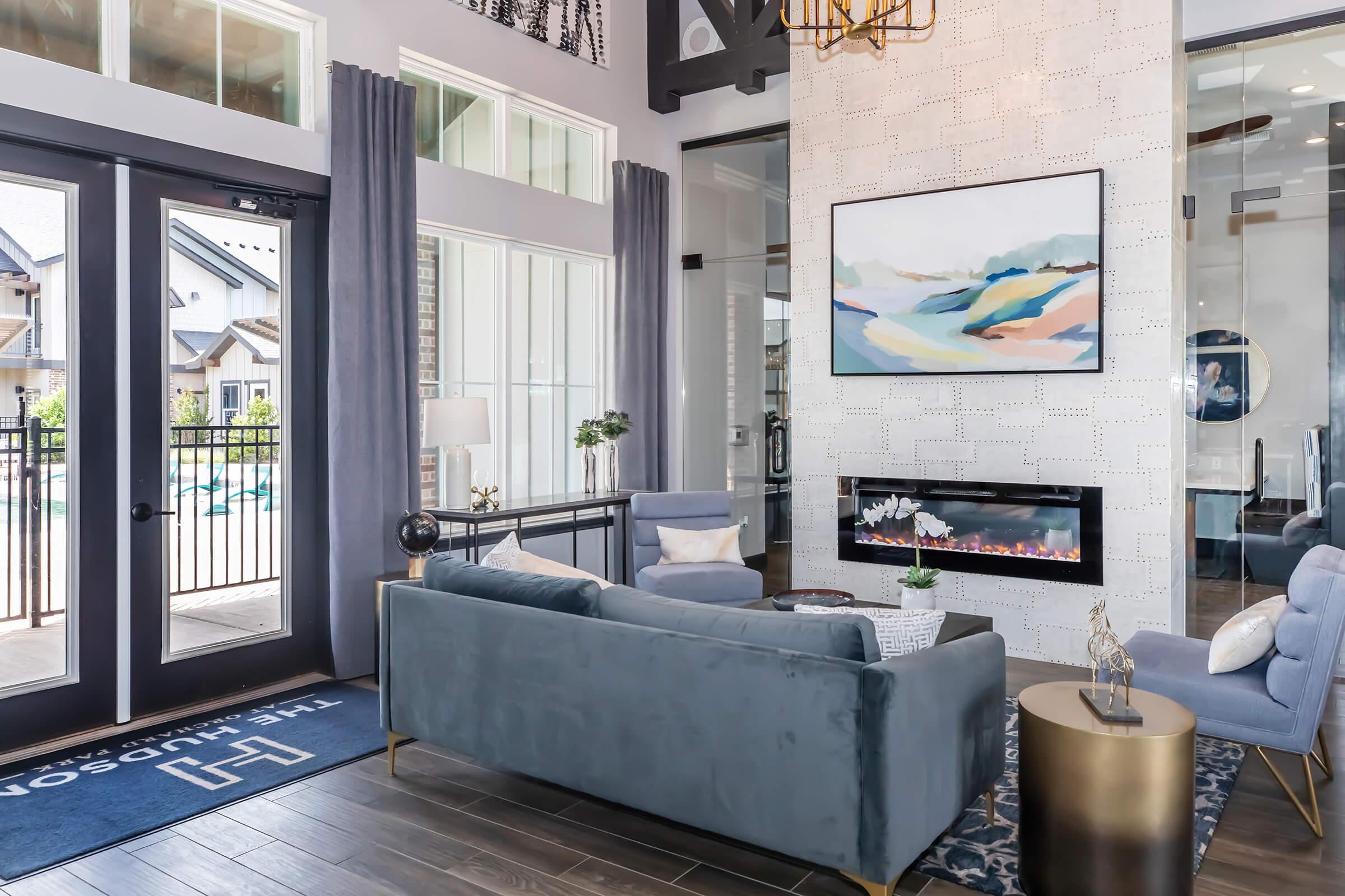 Modern living room with large windows, featuring a stylish gray sofa, armchair, and a gold accent table. A contemporary painting hangs above a sleek electric fireplace. Soft curtains frame the windows, and a decorative rug adds warmth to the space. Outside, a pool area is visible.