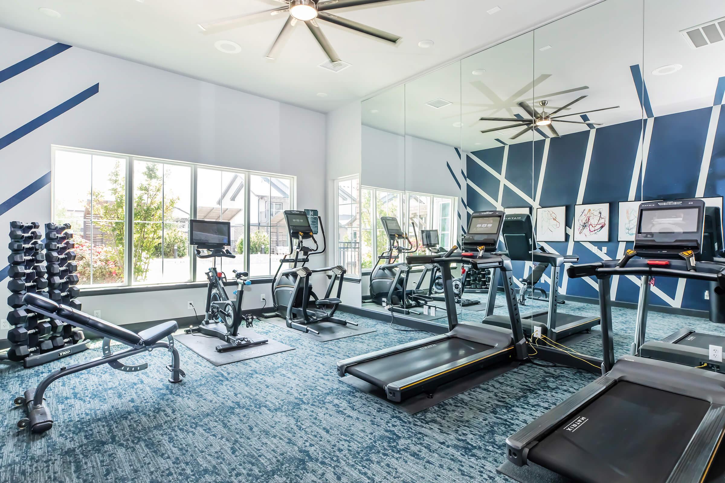A modern gym with cardio machines including treadmills and stationary bikes, a weight bench, and free weights. The walls are accented with blue geometric designs, and large windows allow natural light to fill the space, reflecting in the mirrors. The flooring is a blue carpet, creating a welcoming atmosphere.