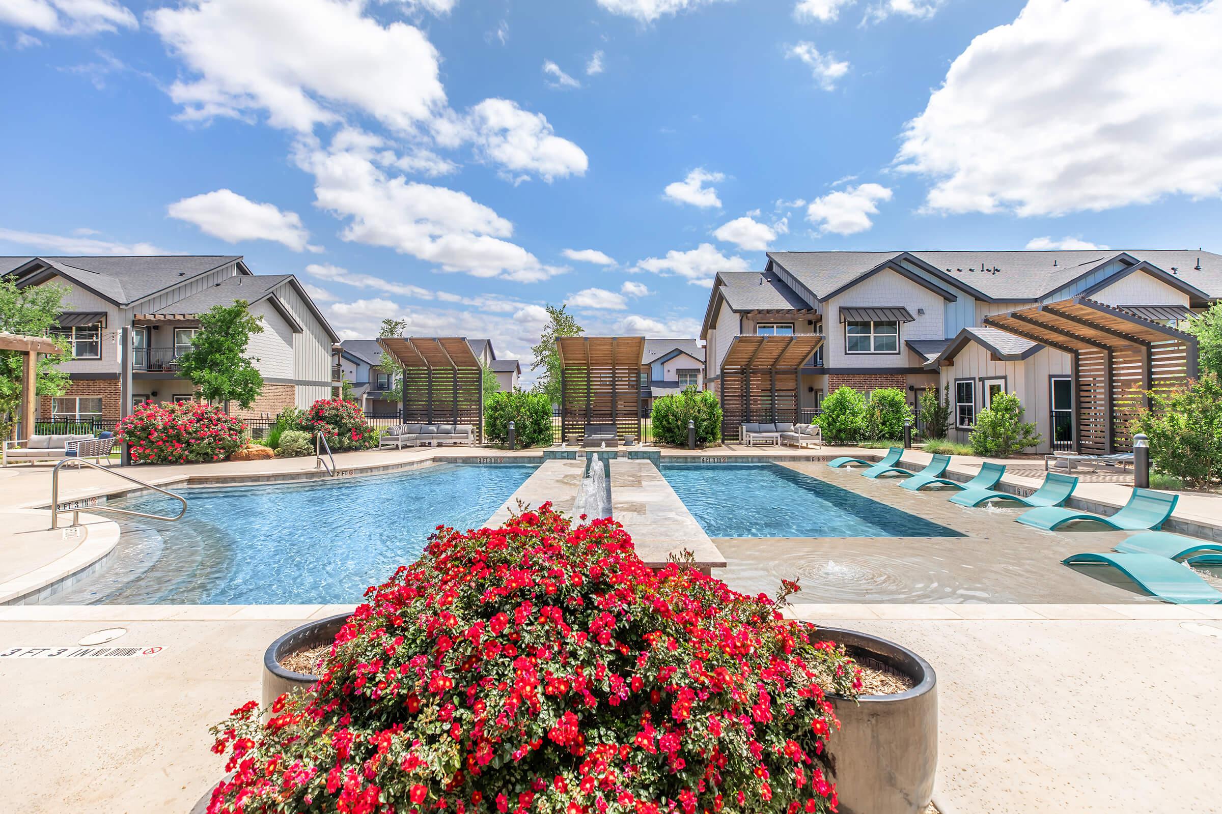 A sunny apartment complex pool area featuring two swimming pools surrounded by lush greenery and colorful flower beds. Lounge chairs are arranged around the pools, and modern buildings can be seen in the background under a partly cloudy sky.