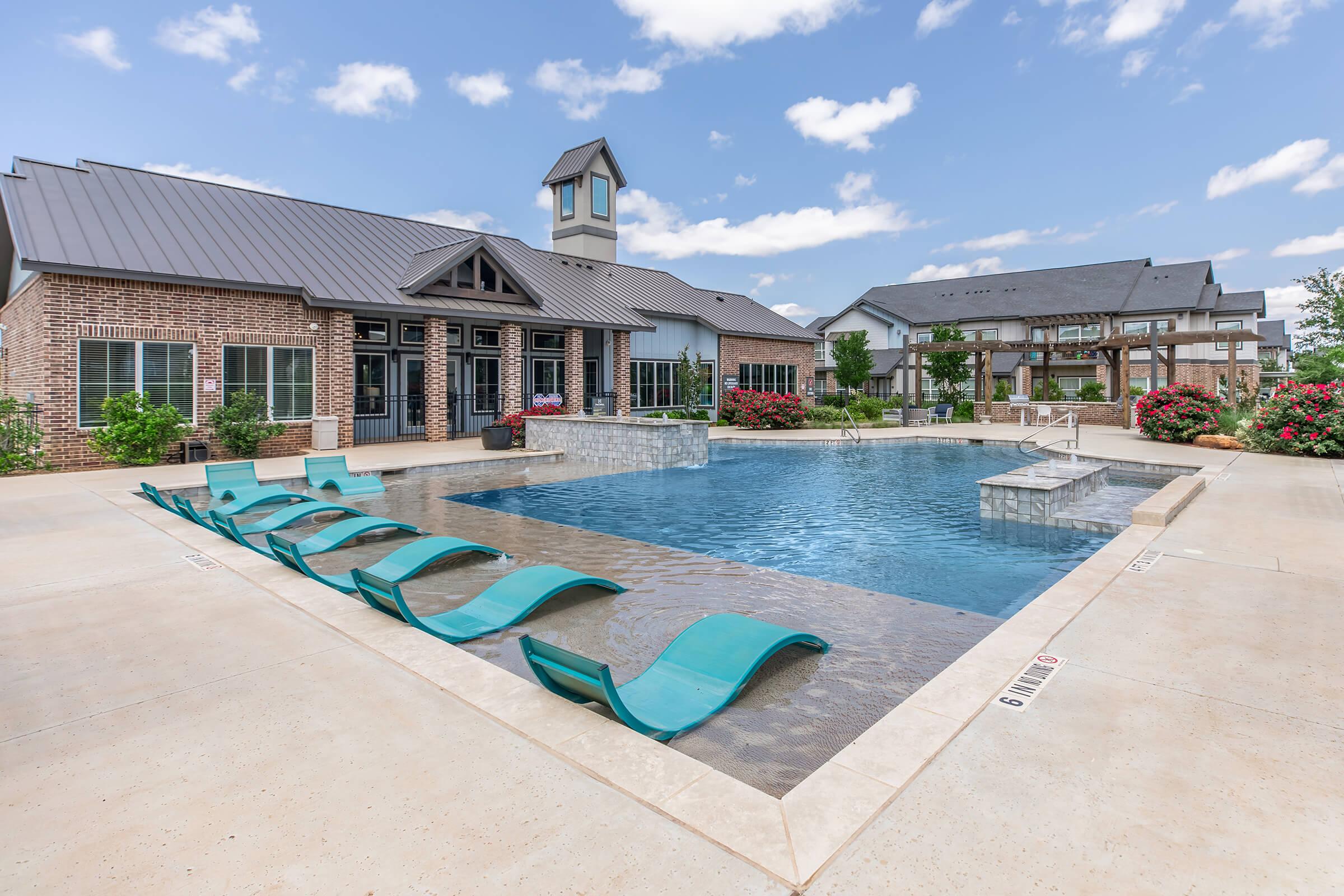 A modern outdoor swimming pool area featuring a clear blue pool with stone accents, surrounded by landscaped gardens and lounge chairs. A building with a peaked roof is visible in the background, adding to the serene atmosphere under a partly cloudy sky.
