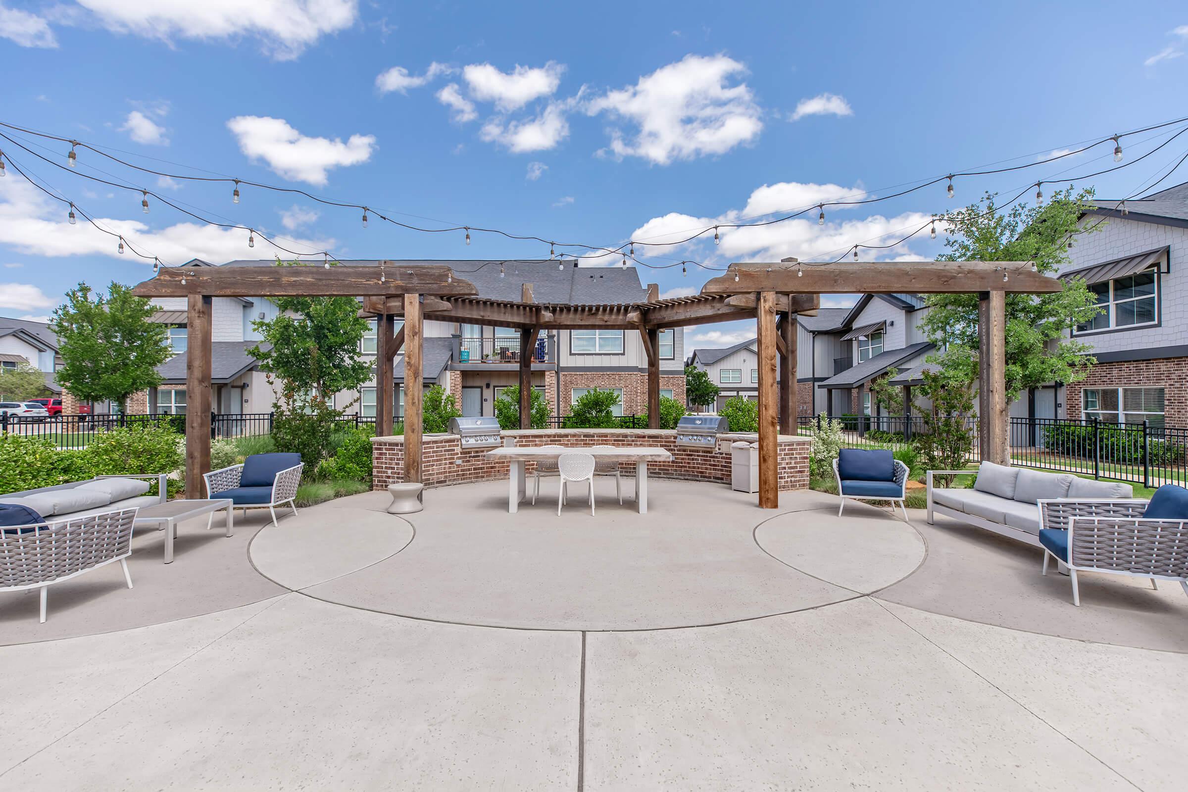 Outdoor patio area with a circular concrete floor, featuring a wooden pergola overhead. Cozy seating options include light-colored sofas and chairs. Surrounding greenery and a residential building in the background, with decorative string lights hanging above, creating a welcoming atmosphere.