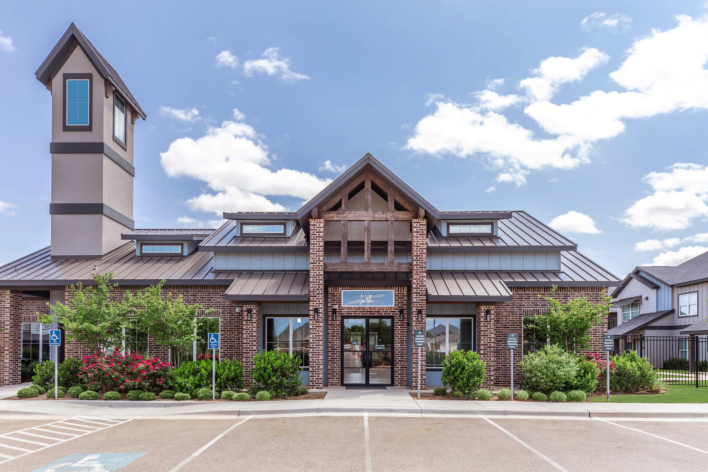 A modern, brick-clad building with a peaked roof and tall clock tower. The front entrance features large glass doors and is flanked by manicured landscaping, including shrubs and small trees. A parking lot is visible in the foreground, with designated accessible parking spaces. The sky is partly cloudy.