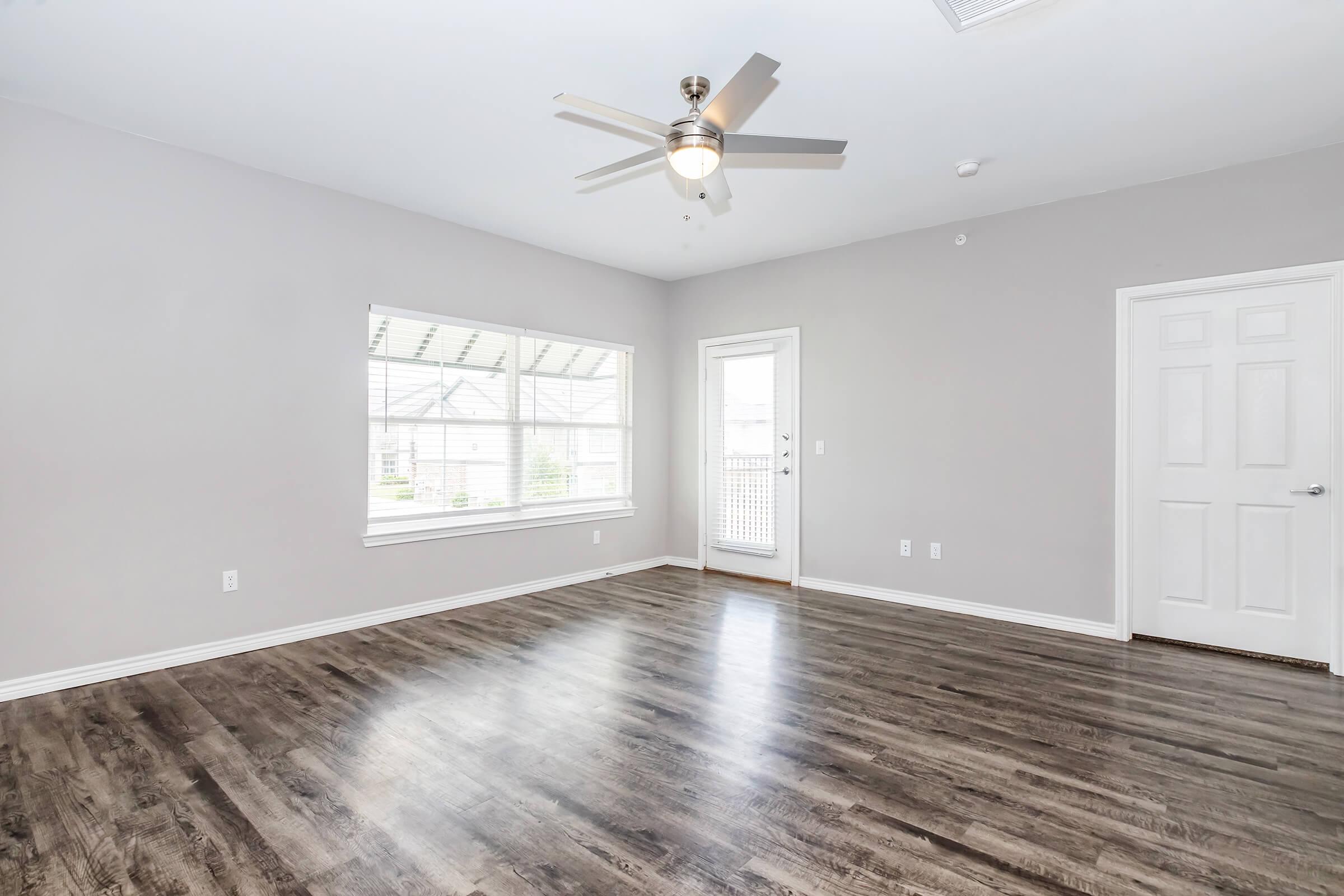 A spacious, empty room featuring light gray walls, a ceiling fan, and large windows allowing natural light. The floor is dark wood, enhancing the modern look. There is a door leading outside and another door on the right side, creating a welcoming atmosphere.