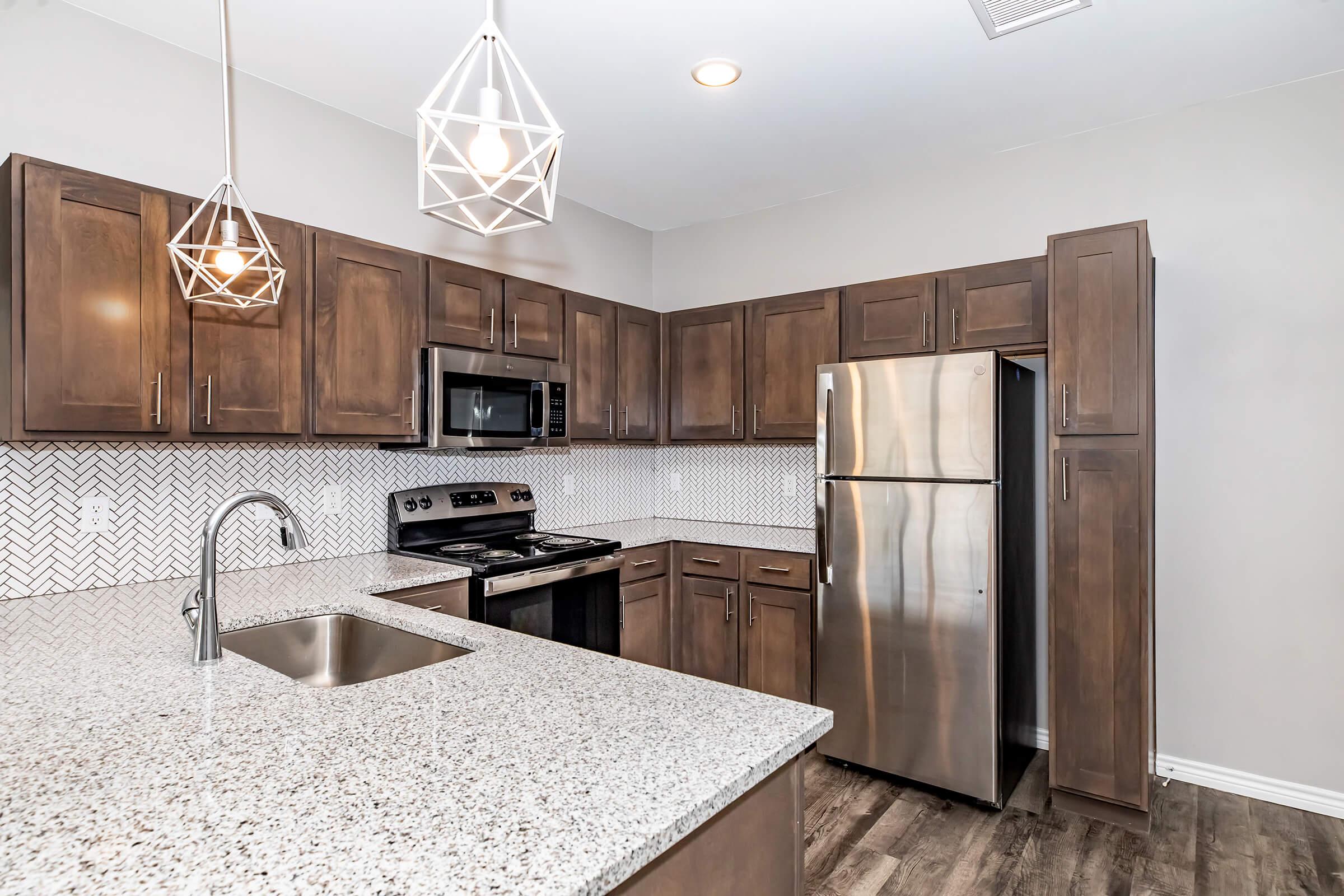 Modern kitchen featuring dark wood cabinets, stainless steel appliances including a refrigerator and oven, a granite countertop with a sink, and stylish hexagonal backsplash tiles. Two pendant lights hang from the ceiling, providing bright illumination in the space.