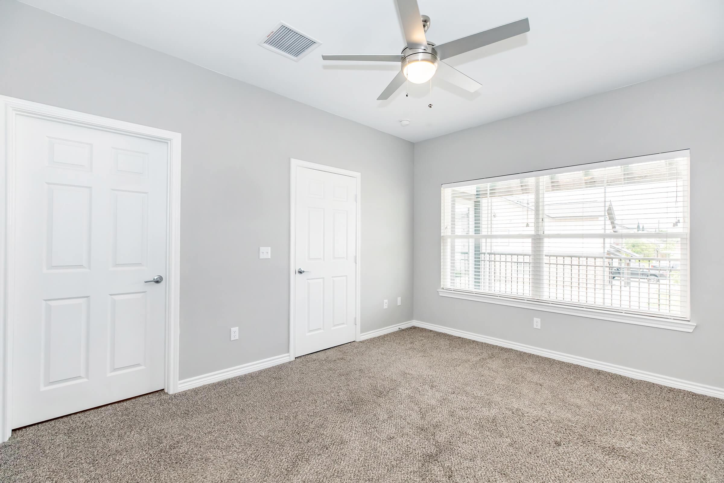 A bright and airy room with light gray walls and carpeted flooring. It features a ceiling fan, two closed doors, and a large window with blinds that offers natural light. The room is empty, providing a spacious feel perfect for various uses.