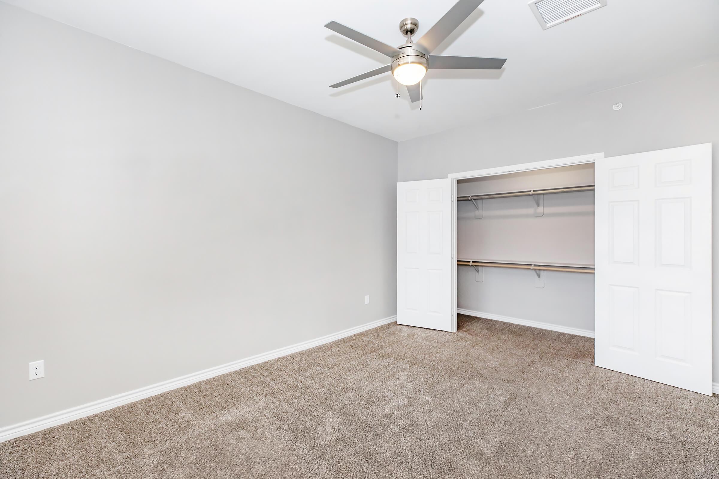 Empty room with gray walls and carpet. It features a ceiling fan, a closet with double doors, and no furniture. The space has good natural light, creating a spacious feel.