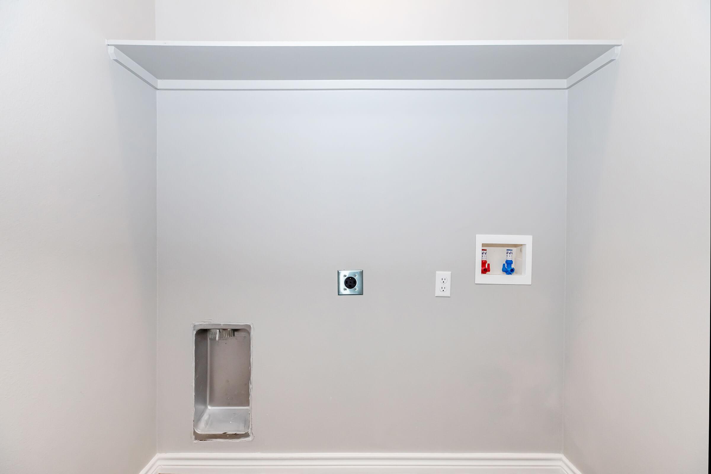 A blank wall in a room featuring a built-in shelf at the top. On the wall, there is a rectangular cutout at the left, a circular electrical outlet, and a dual water connection on the right, suggesting it may be intended for laundry appliances or similar fixtures. The wall is painted a light gray.