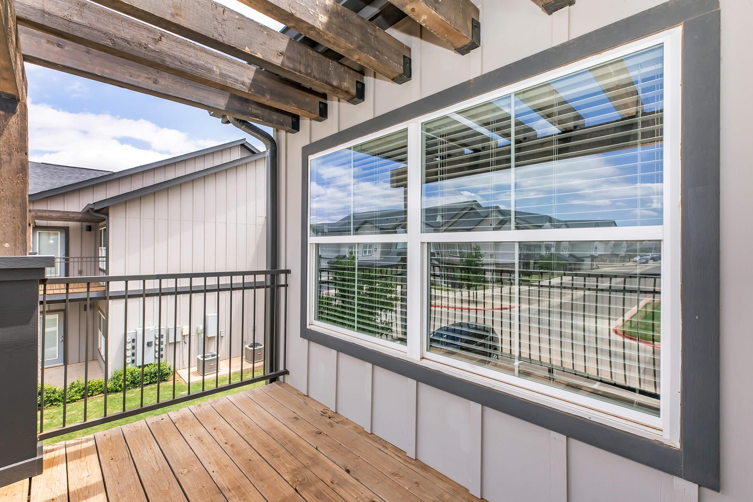 View of a balcony with wooden flooring and a railing, featuring large windows reflecting the blue sky and nearby buildings. The surrounding architecture includes light-colored exteriors and green landscaping in the background.
