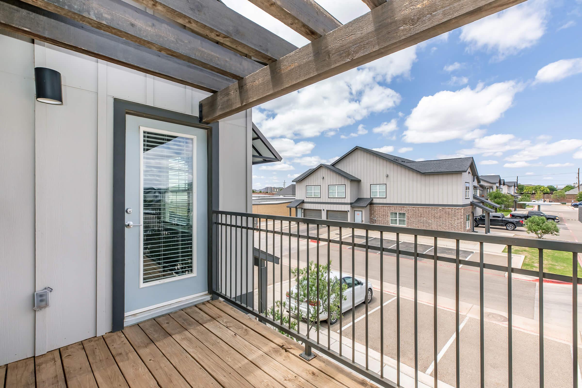 A wooden balcony with a metal railing, leading to a glass door with blinds. The view showcases neighboring buildings under a partly cloudy sky. The setting features a relaxed, residential atmosphere.