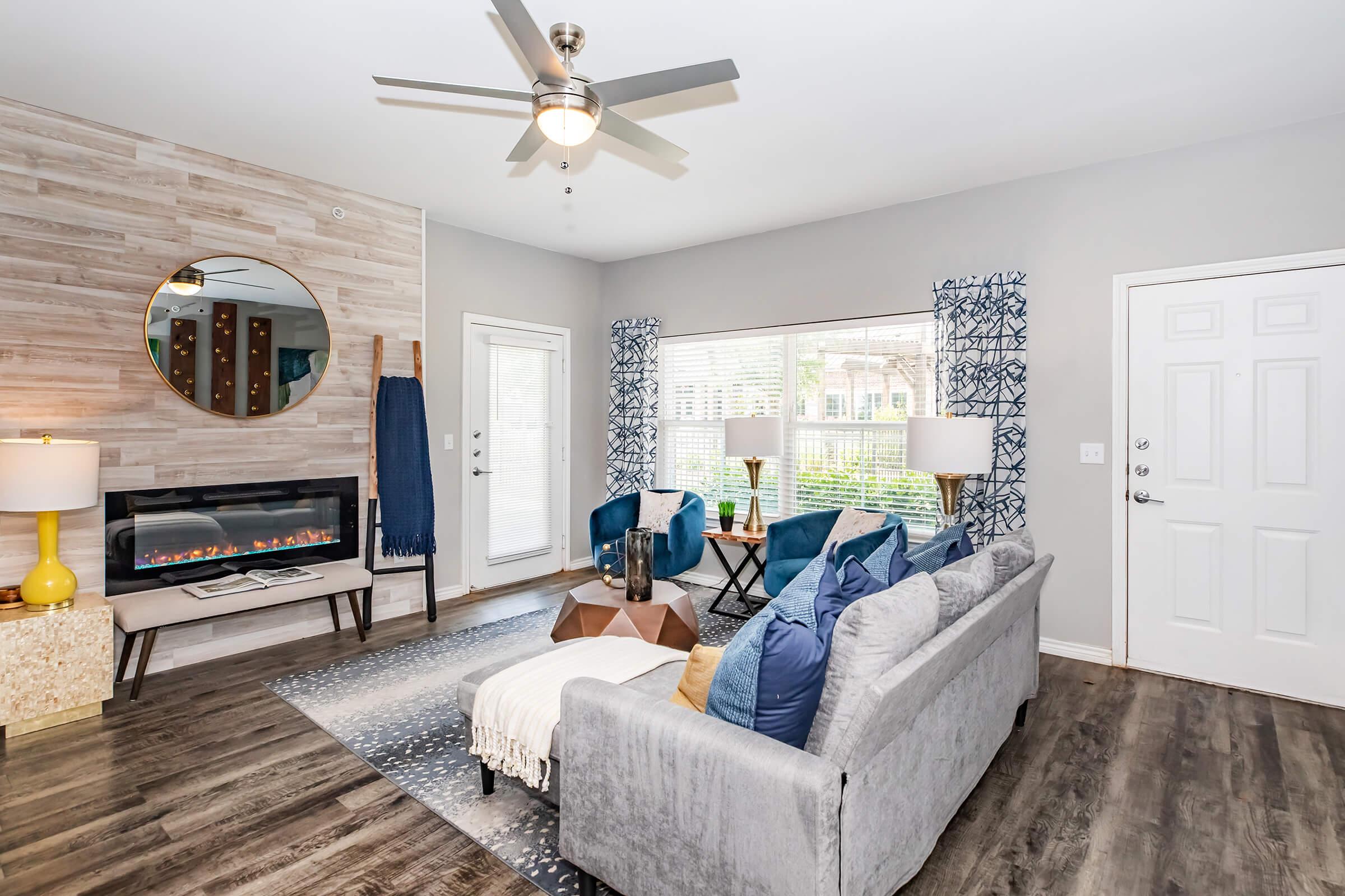 Cozy living room featuring a gray sofa with decorative pillows, blue armchairs, a circular mirror, and a modern electric fireplace. Large windows allow natural light, complemented by wooden flooring and contemporary decor. A stylish ceiling fan enhances the comfortable atmosphere.