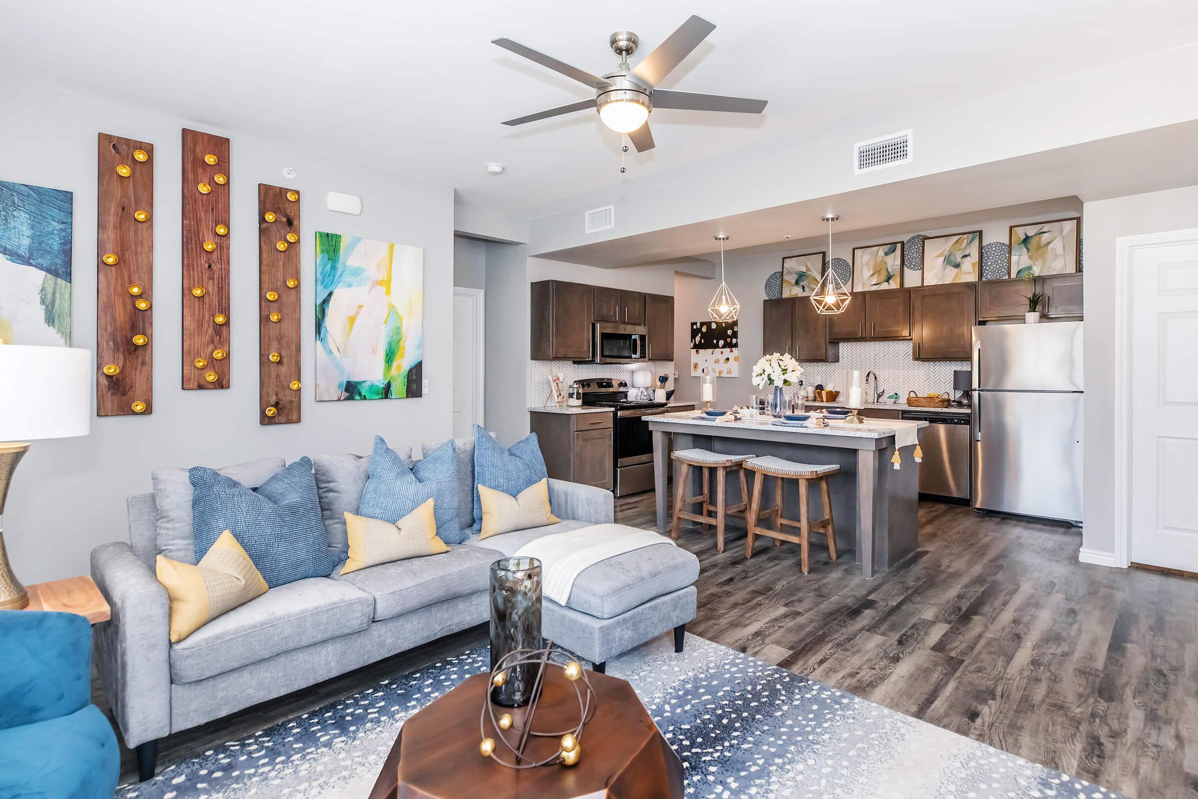 A modern living room and kitchen area featuring a gray sectional sofa with blue and yellow pillows, wooden wall decor, and a stylish gray rug. The kitchen includes dark cabinets, stainless steel appliances, and a central island with bar stools. Bright artwork enhances the contemporary decor.